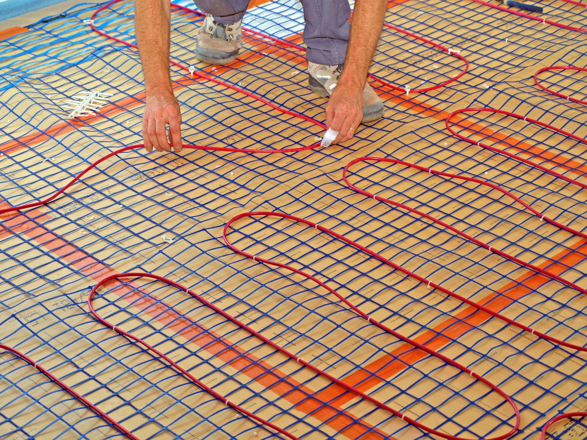 Personne installant des câbles chauffants rouges sur un plancher en treillis bleu, en préparation de la pose du revêtement de sol.