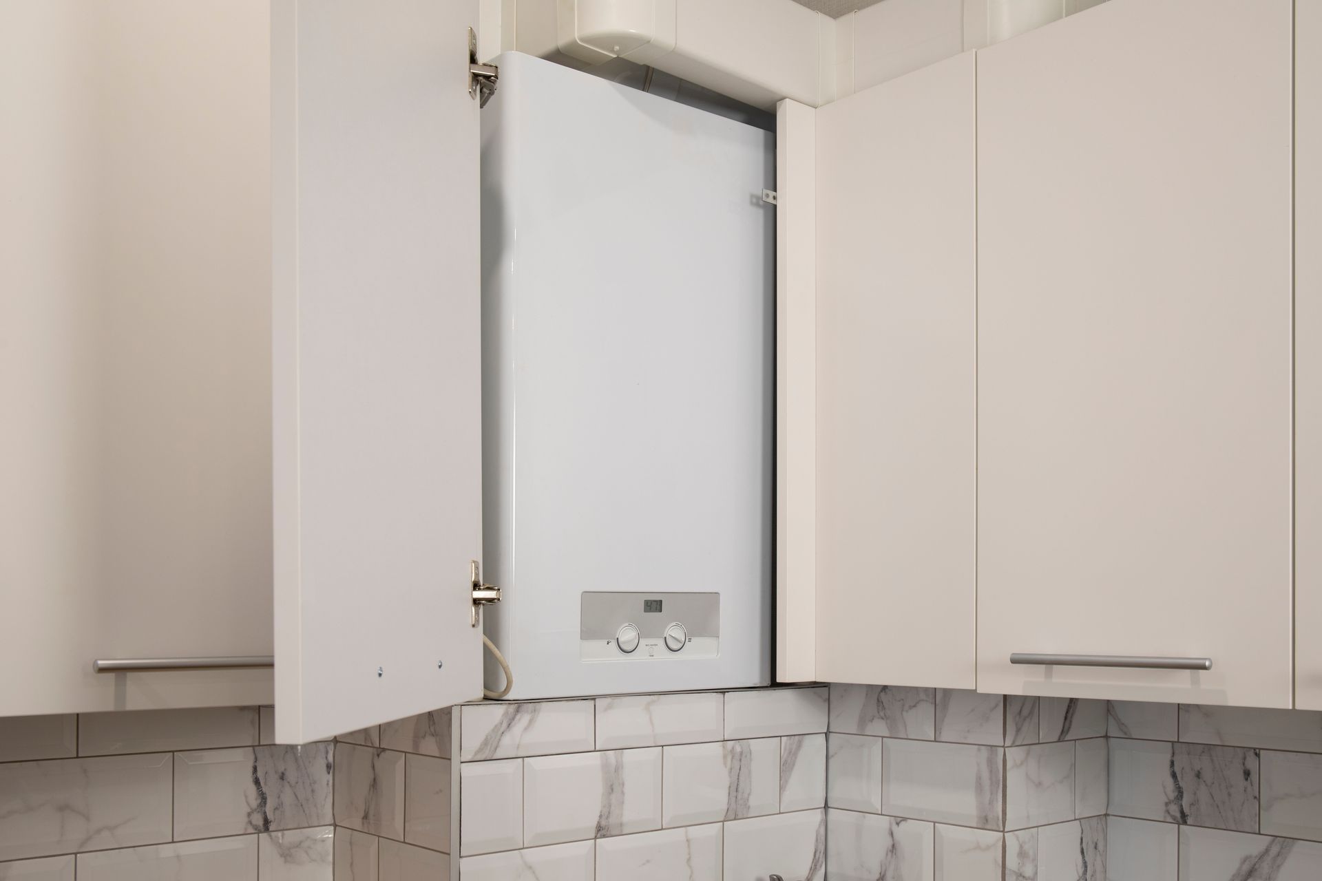 Chaudière murale blanche dans un meuble de cuisine, une porte ouverte. Crédence en marbre clair.