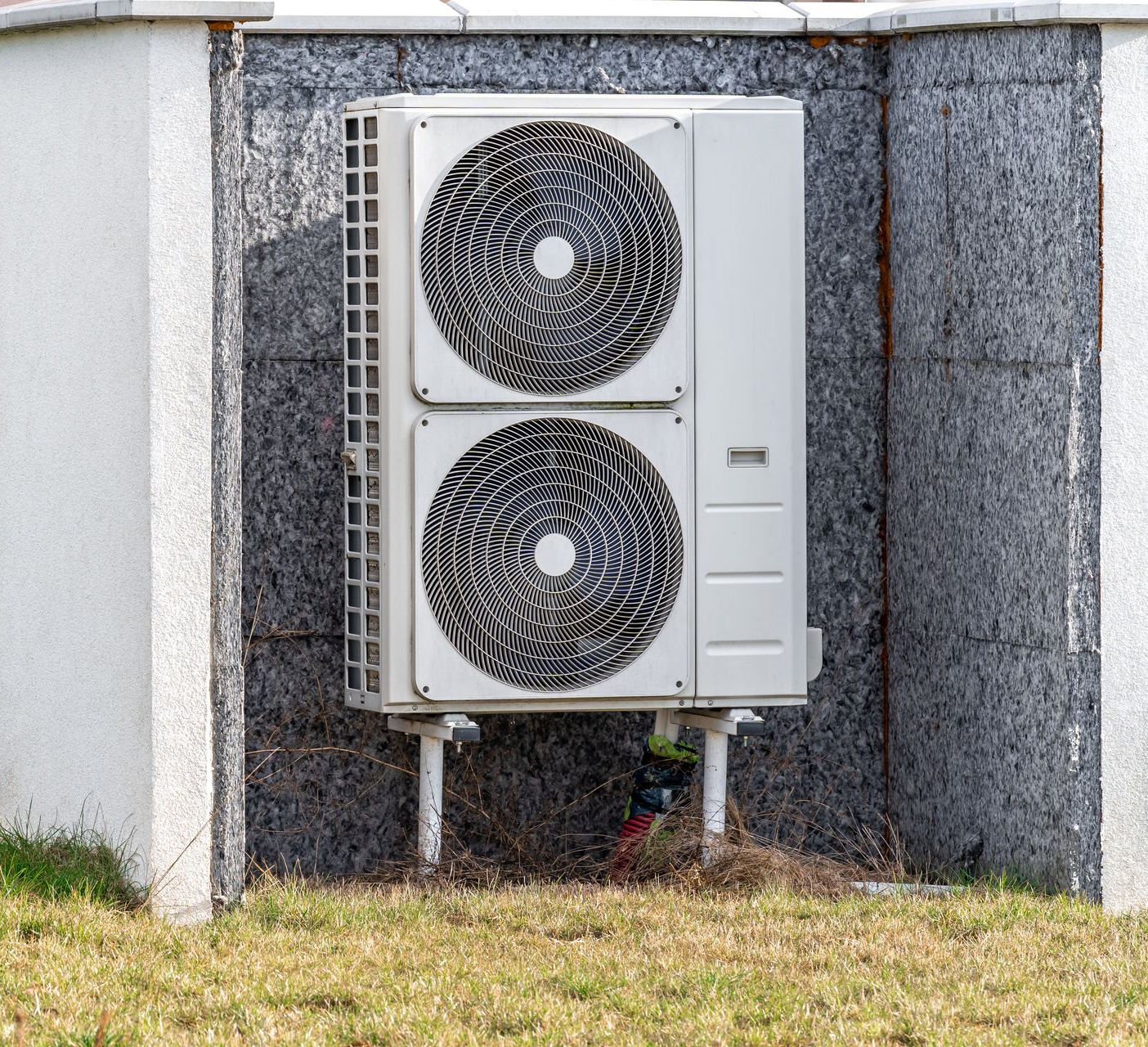 Unité de climatisation extérieure, boîtier rectangulaire de couleur claire avec deux grilles d'aération circulaires, montée sur pieds près d'un bâtiment.