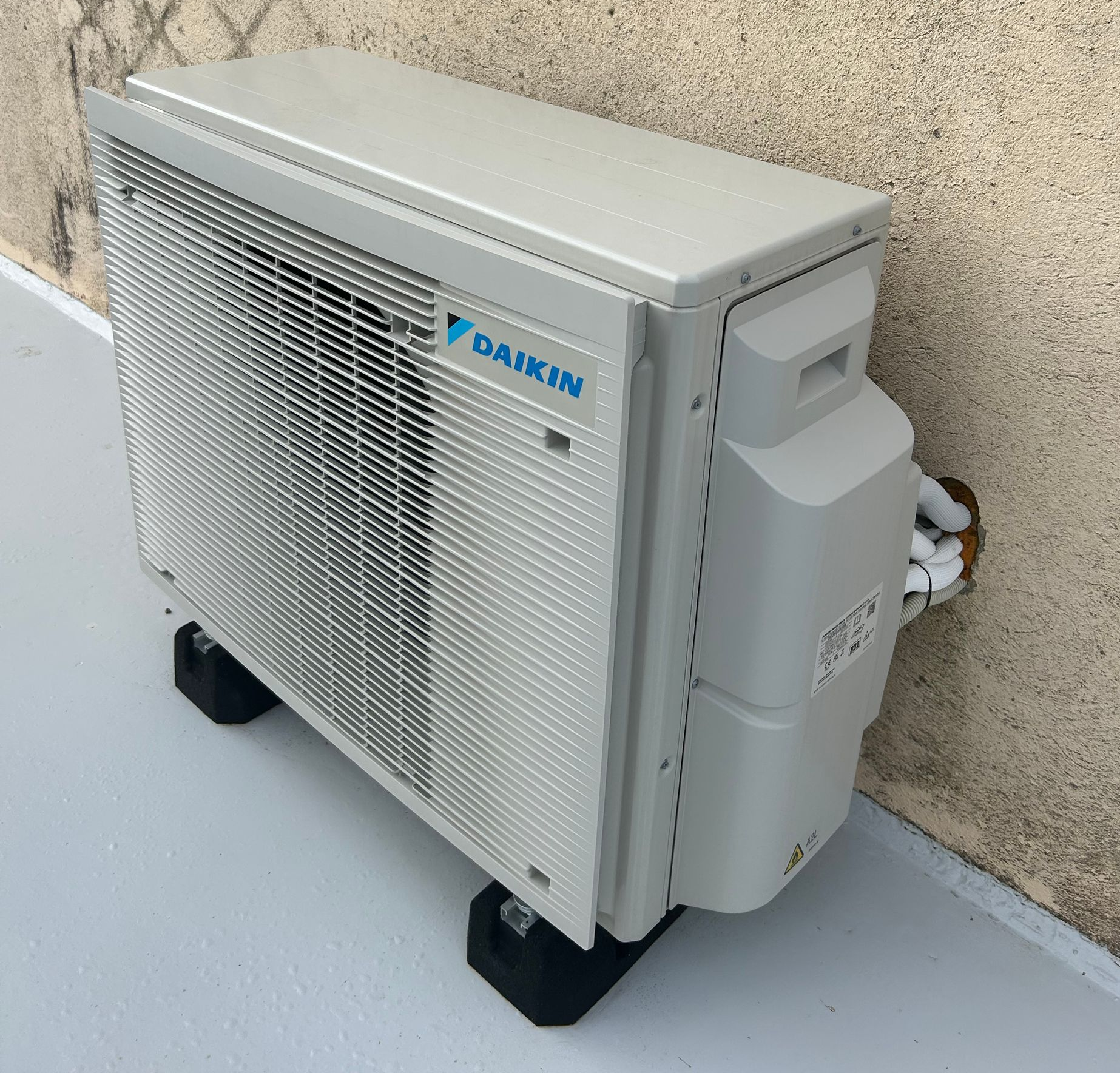 Unité de climatisation Daikin sur blocs noirs, fixée à l'extérieur contre un mur beige, sur une surface peinte en gris.