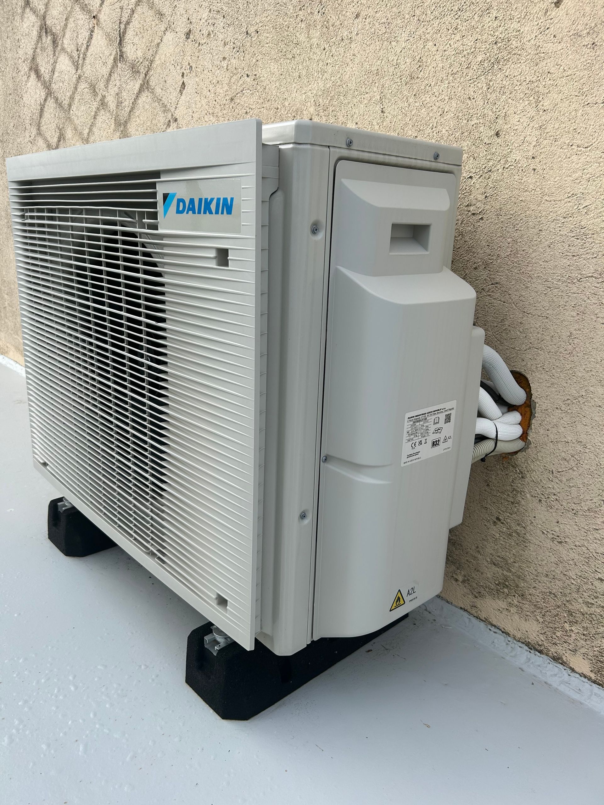 Unité de climatisation Daikin sur blocs noirs, fixée sur une surface gris clair, contre un mur brun clair.