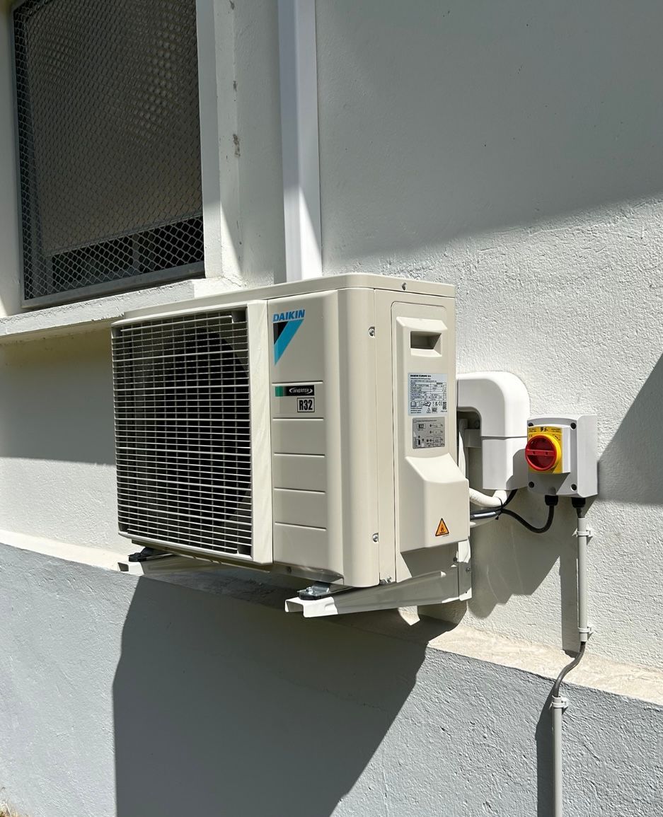 Unité de climatisation extérieure fixée sur un mur blanc du bâtiment, avec un boîtier électrique.