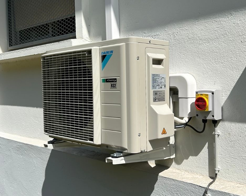 Un climatiseur fixé sur la façade blanche d'un bâtiment, avec un interrupteur rouge à côté.