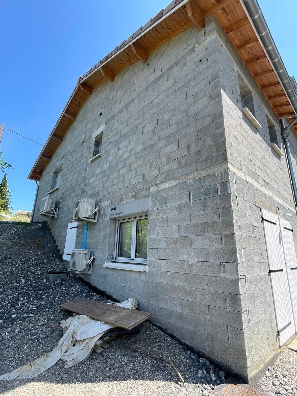 Maison en parpaings gris sur une pente, avec toit en pente et fenêtres, vue d'en bas