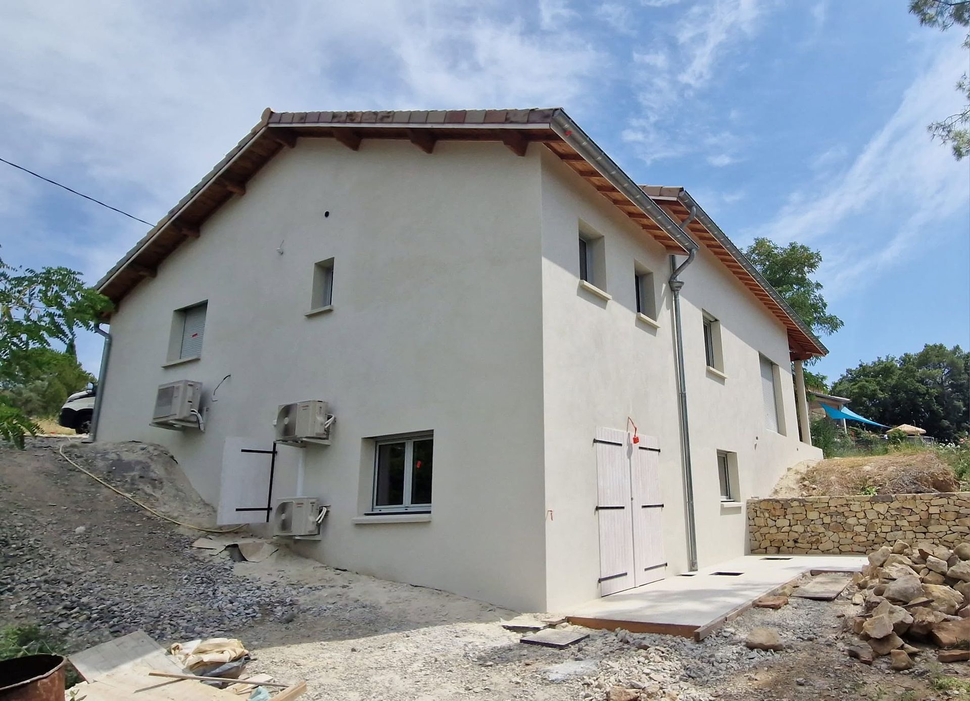 Extérieur d'une maison blanche avec un toit en pente, des climatiseurs et un aménagement paysager en gravier sous un ciel bleu