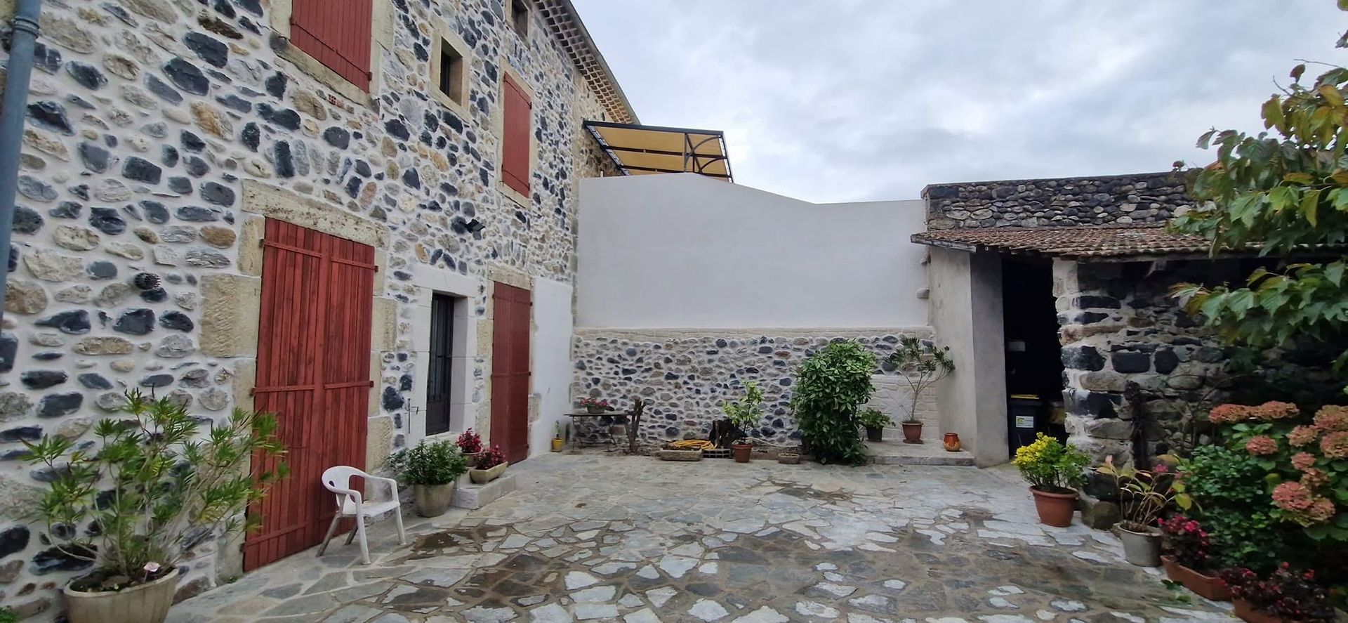 Cour en pierre entre des bâtiments rustiques aux murs de pierre, portes rouges et des plantes en pot sous un ciel nuageux