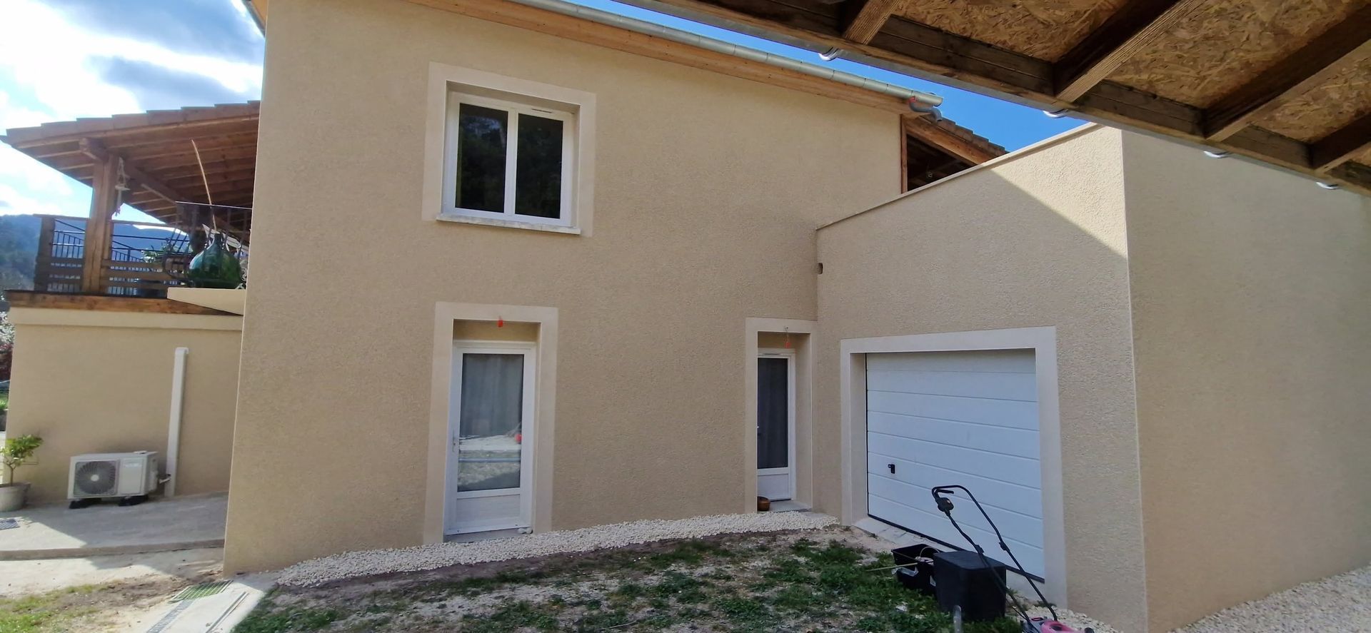 Maison à l'extérieur en stuc beige, avec des fenêtres à encadrement blanc, une porte de garage et un petit chemin de pierre.