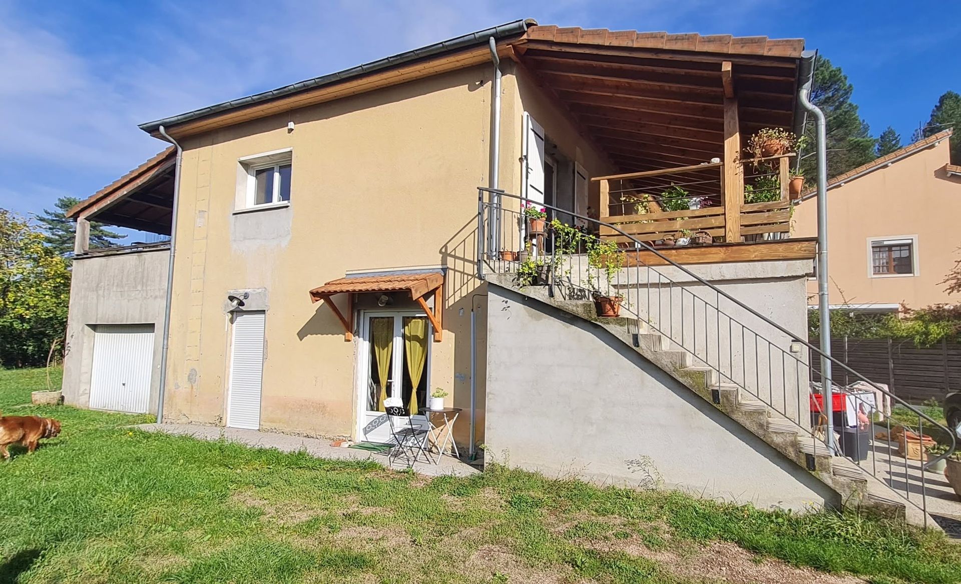 Maison beige à deux étages avec un escalier extérieur, un balcon et un jardin gazonné baigné de soleil.