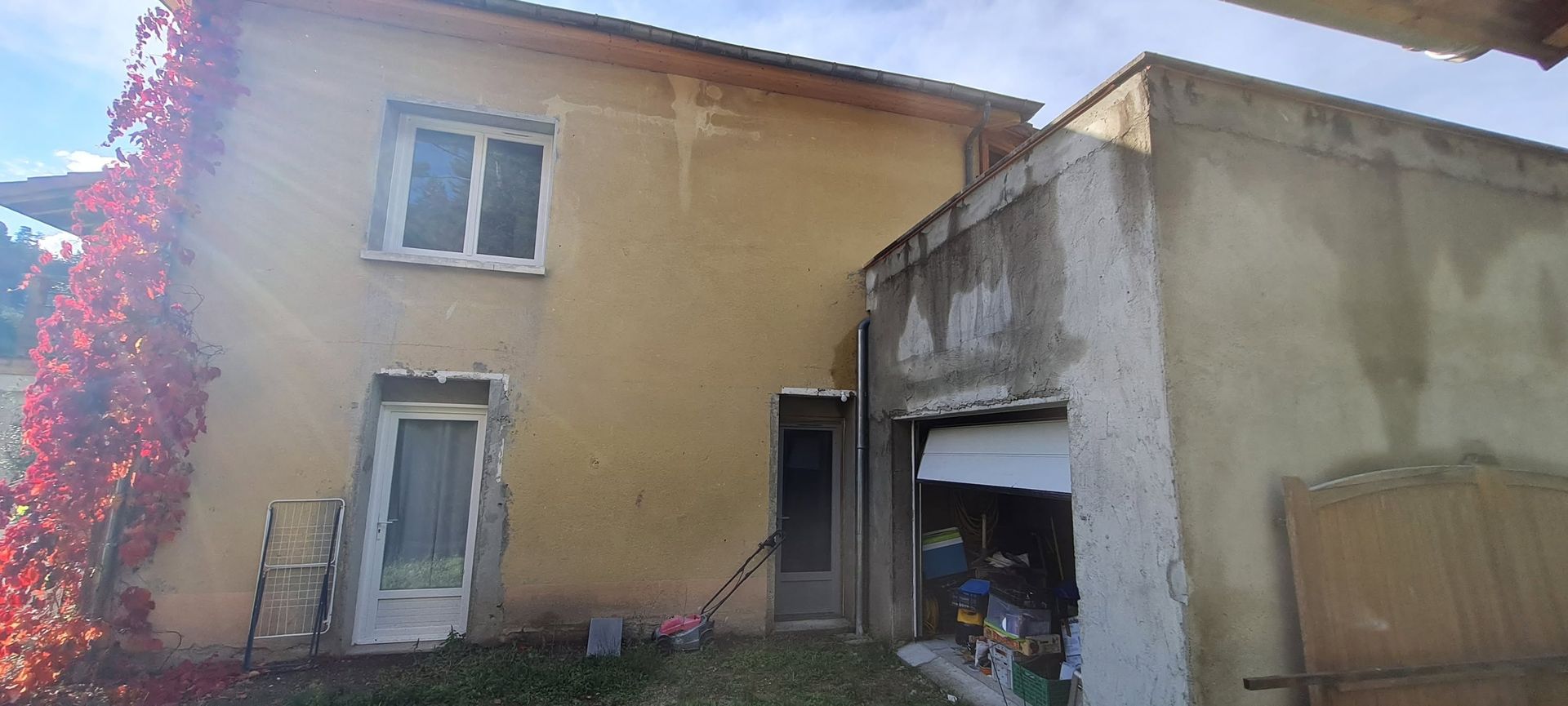 Maison jaune à deux étages avec une extension grise défraîchie et une vigne fleurie rose sur la gauche.