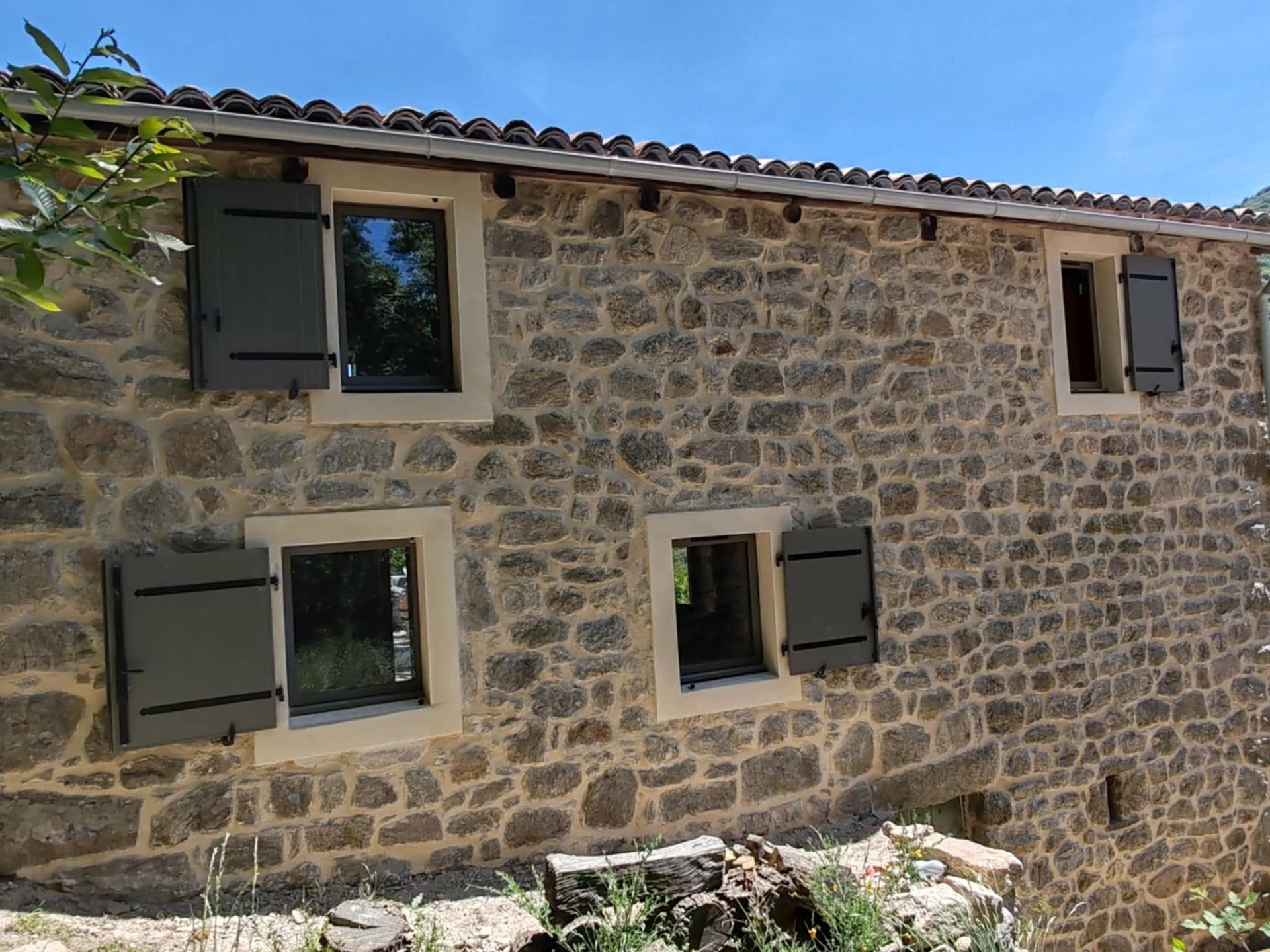 Mur d'une maison en pierre aux volets sombres sous un ciel bleu, vu de l'extérieur