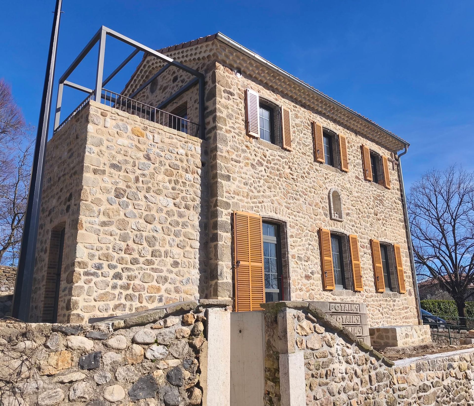 Maison en pierre aux volets en bois, sous un ciel bleu, vue d'en bas, derrière un muret en pierre.