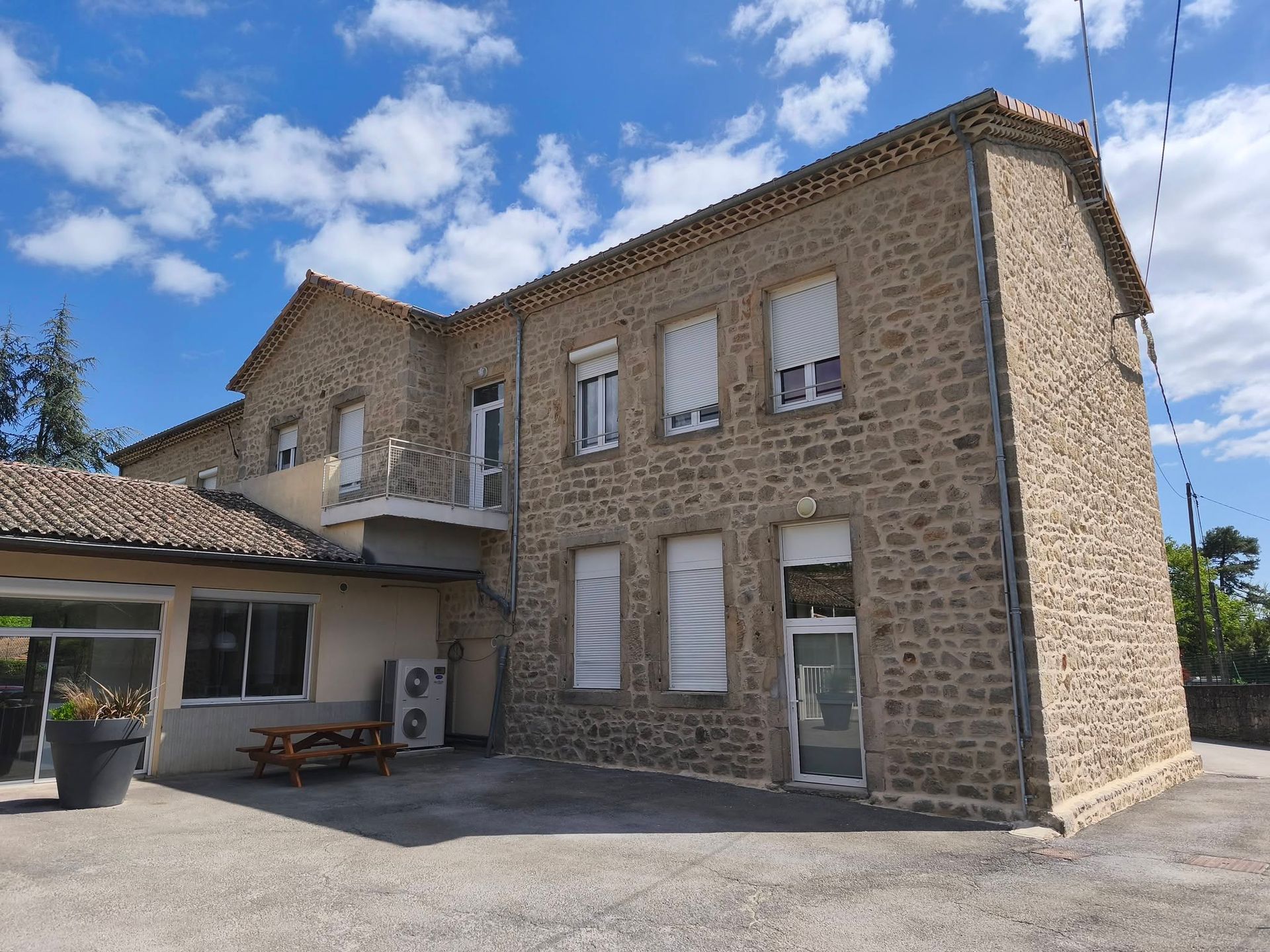 Maison en pierre avec un toit de tuiles, des stores blancs et un petit patio couvert sous un ciel bleu.