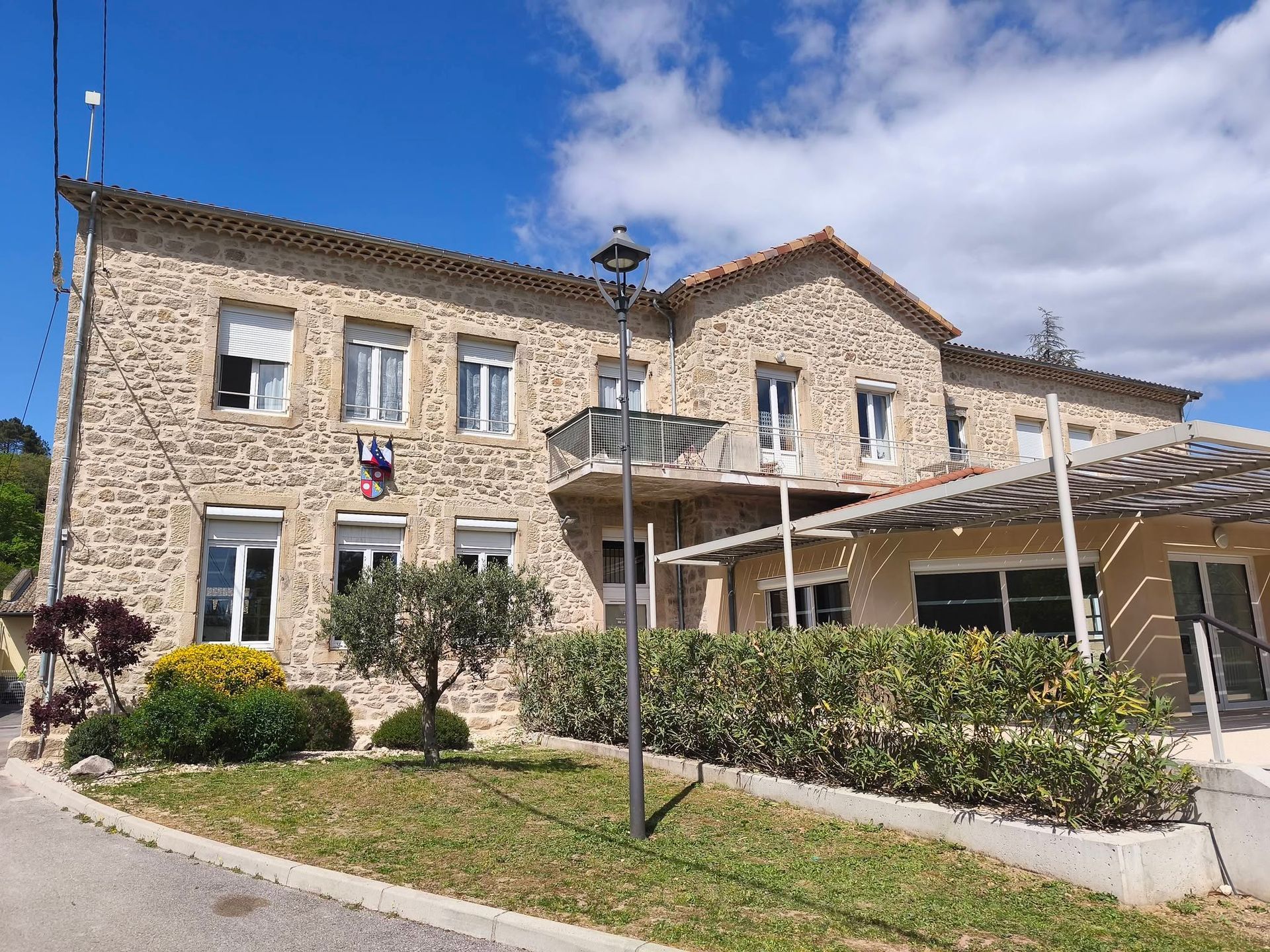 Bâtiment en pierre avec une entrée couverte, un drapeau et un parterres de fleurs par une journée ensoleillée
