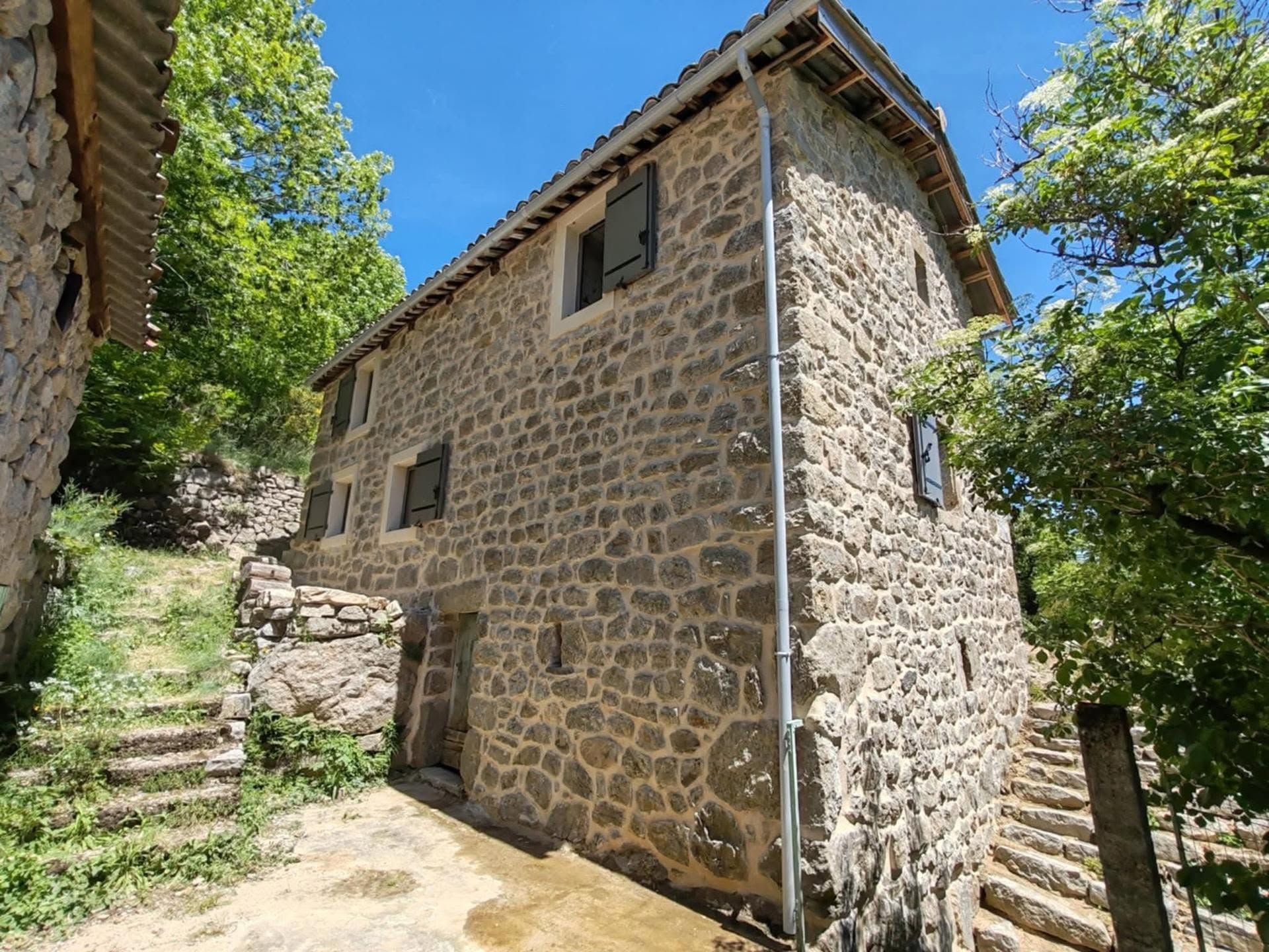 Maison en pierre à flanc de colline, au bout d'un escalier de pierre, entourée d'arbres verdoyants sous un ciel bleu.