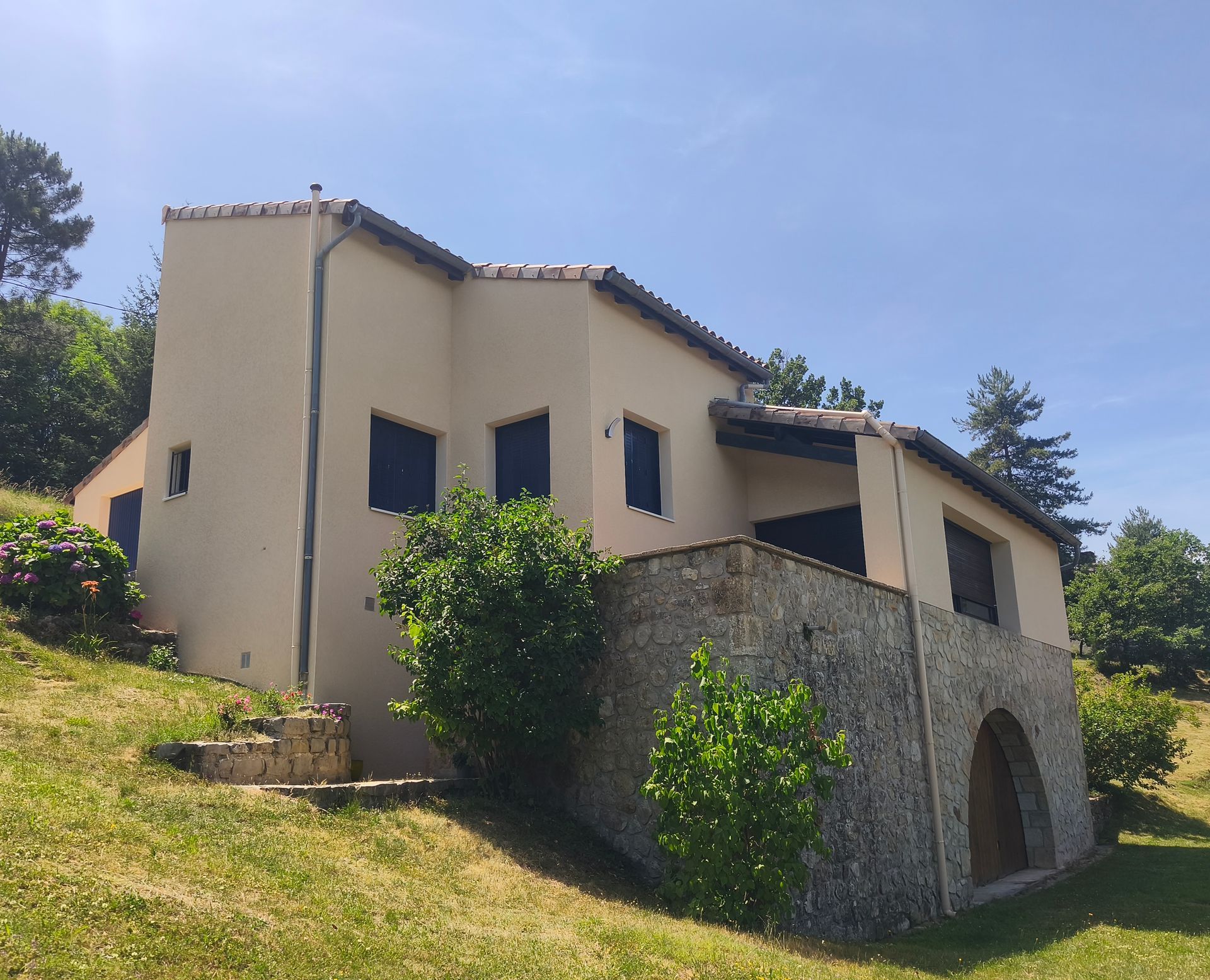 Maison en pierre aux murs beiges et aux fenêtres sombres, sur un coteau herbeux sous un ciel d'un bleu limpide.