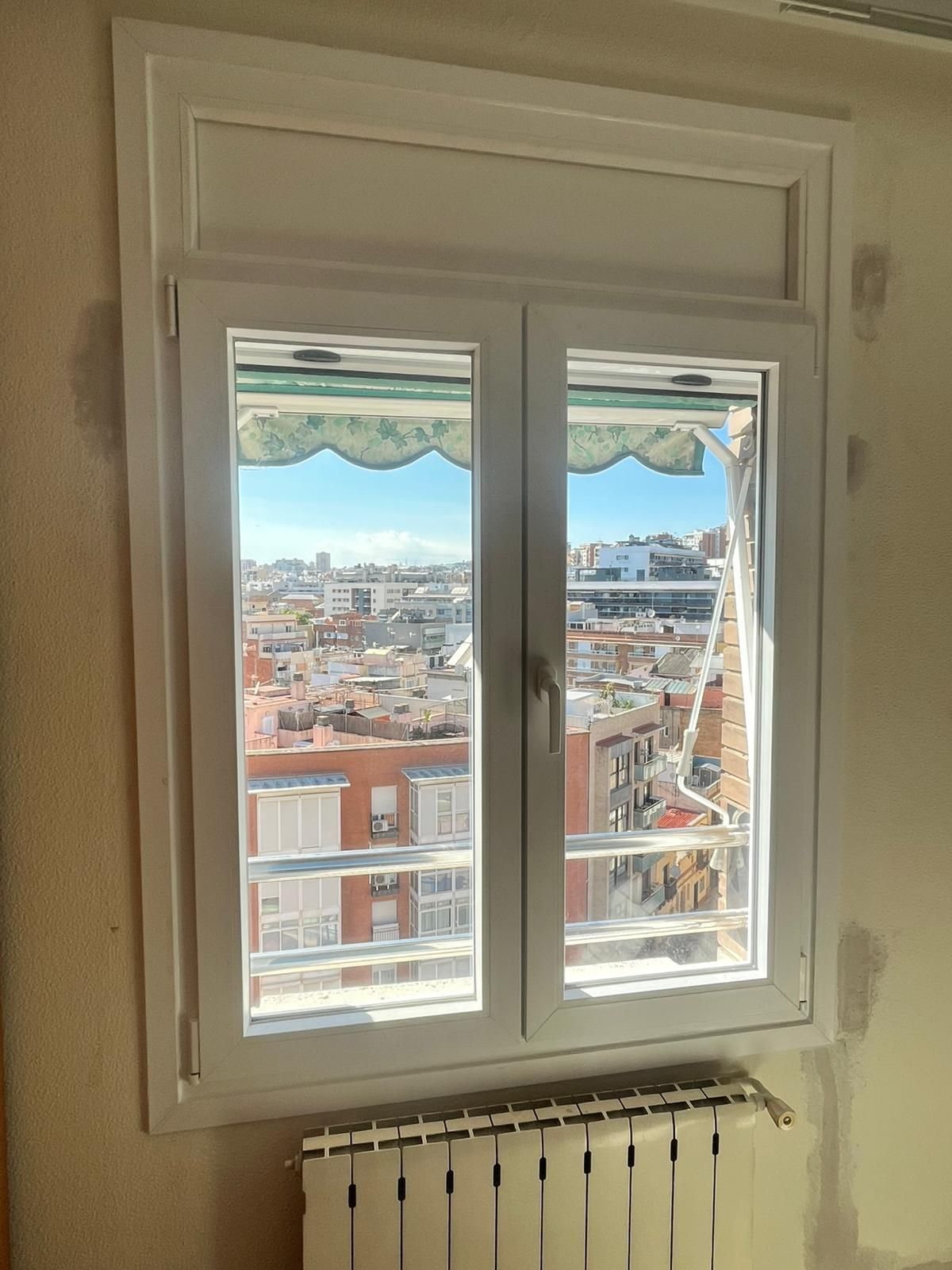 Ventana blanca con balcón con vistas a un paisaje urbano soleado y toldo a rayas, sobre un radiador.