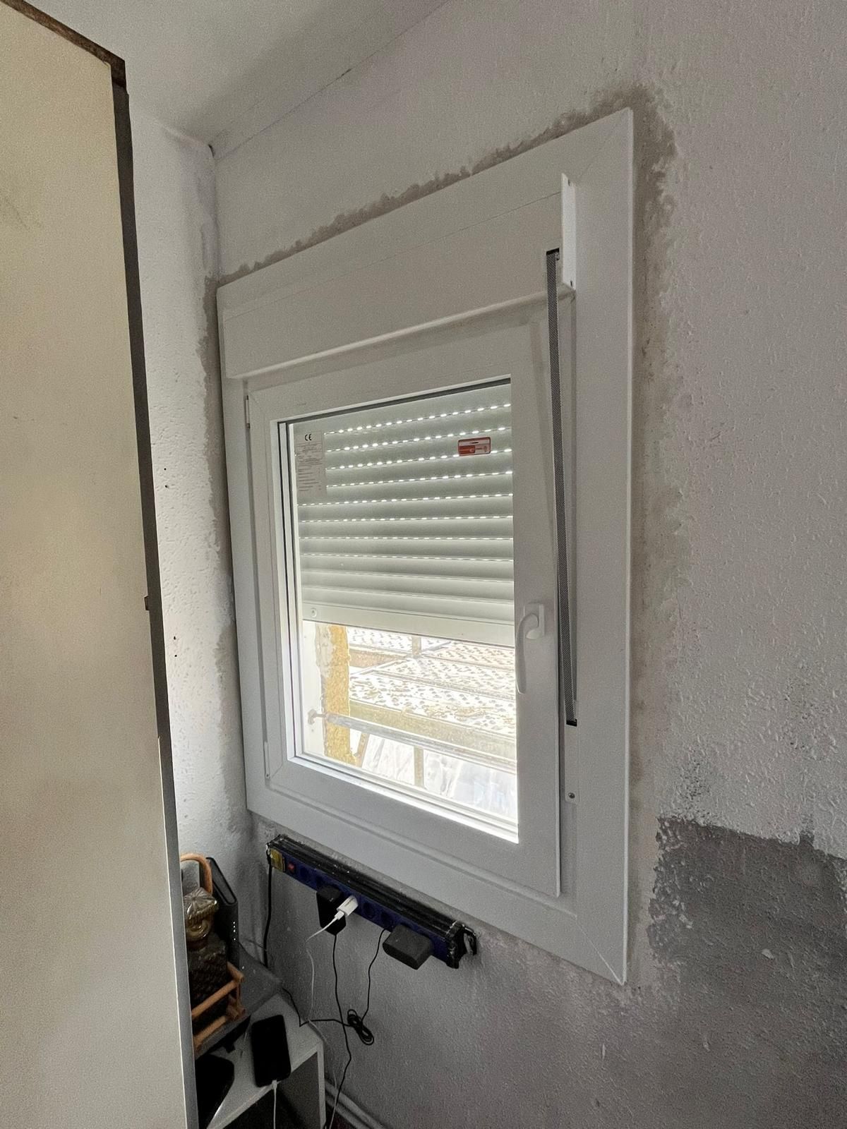 Ventana blanca con persiana enrollable en una habitación vacía, con herramientas y una pared sin terminar debajo.