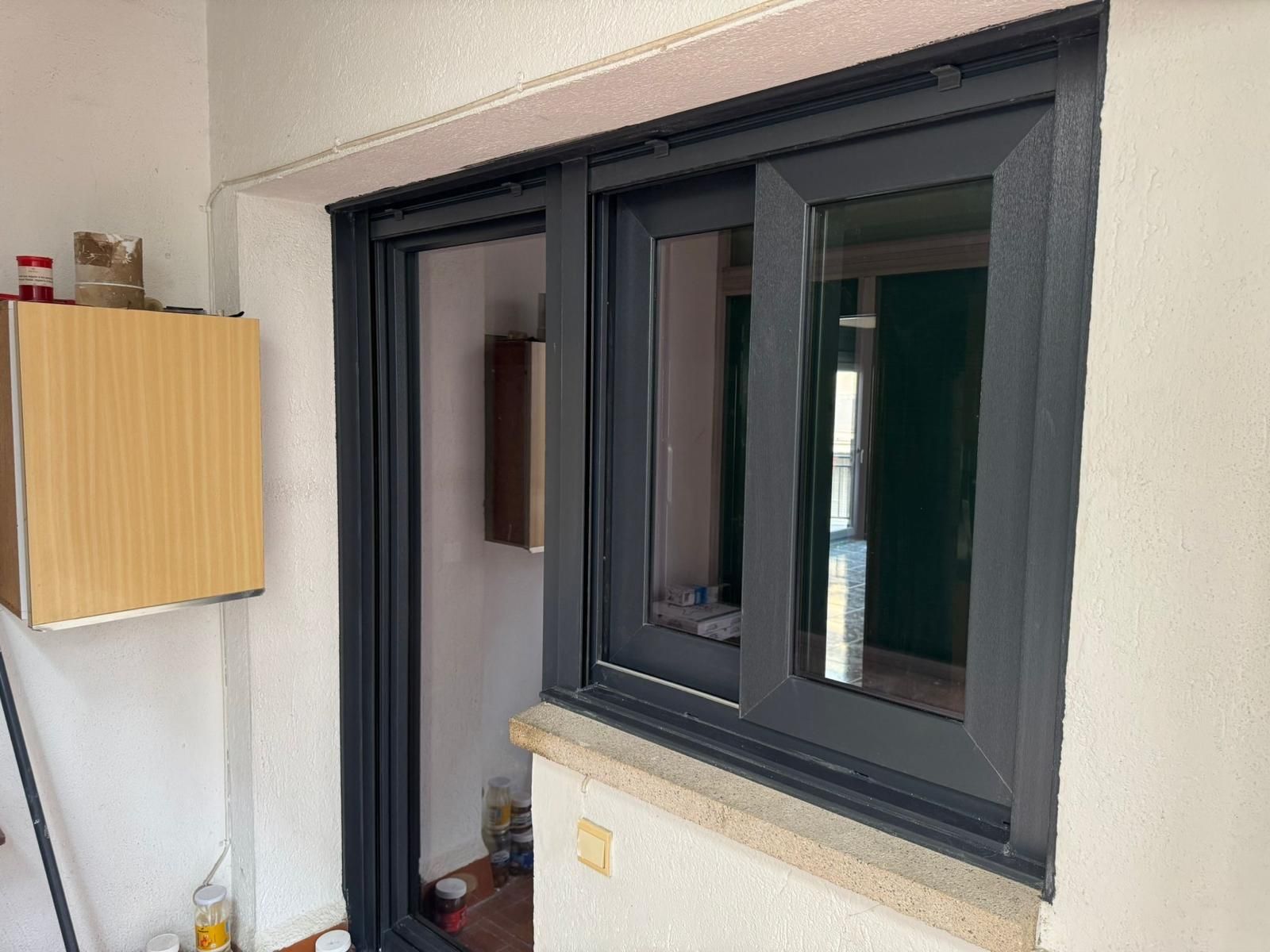 Ventana interior con marco negro y un armario de pared de madera en una habitación blanca.