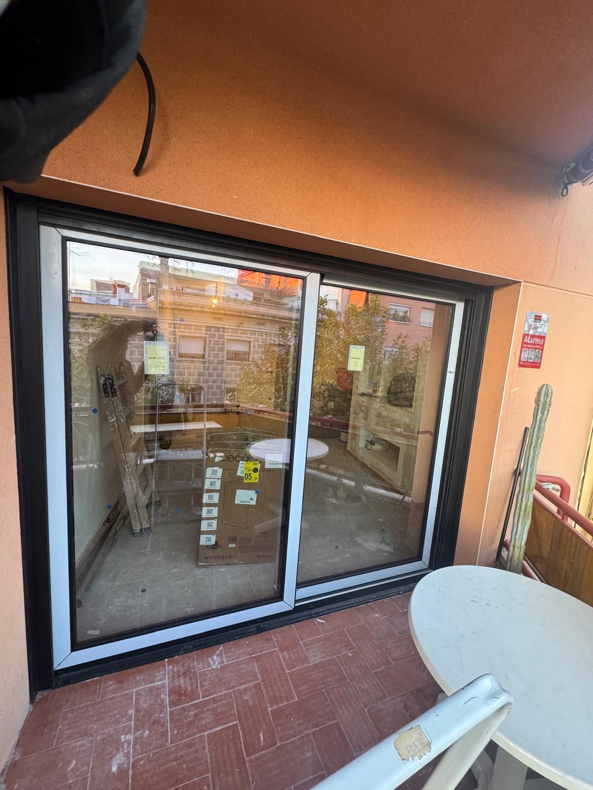 Una puerta corredera de cristal junto a una mesa redonda blanca en un balcón con suelo de baldosas de terracota.