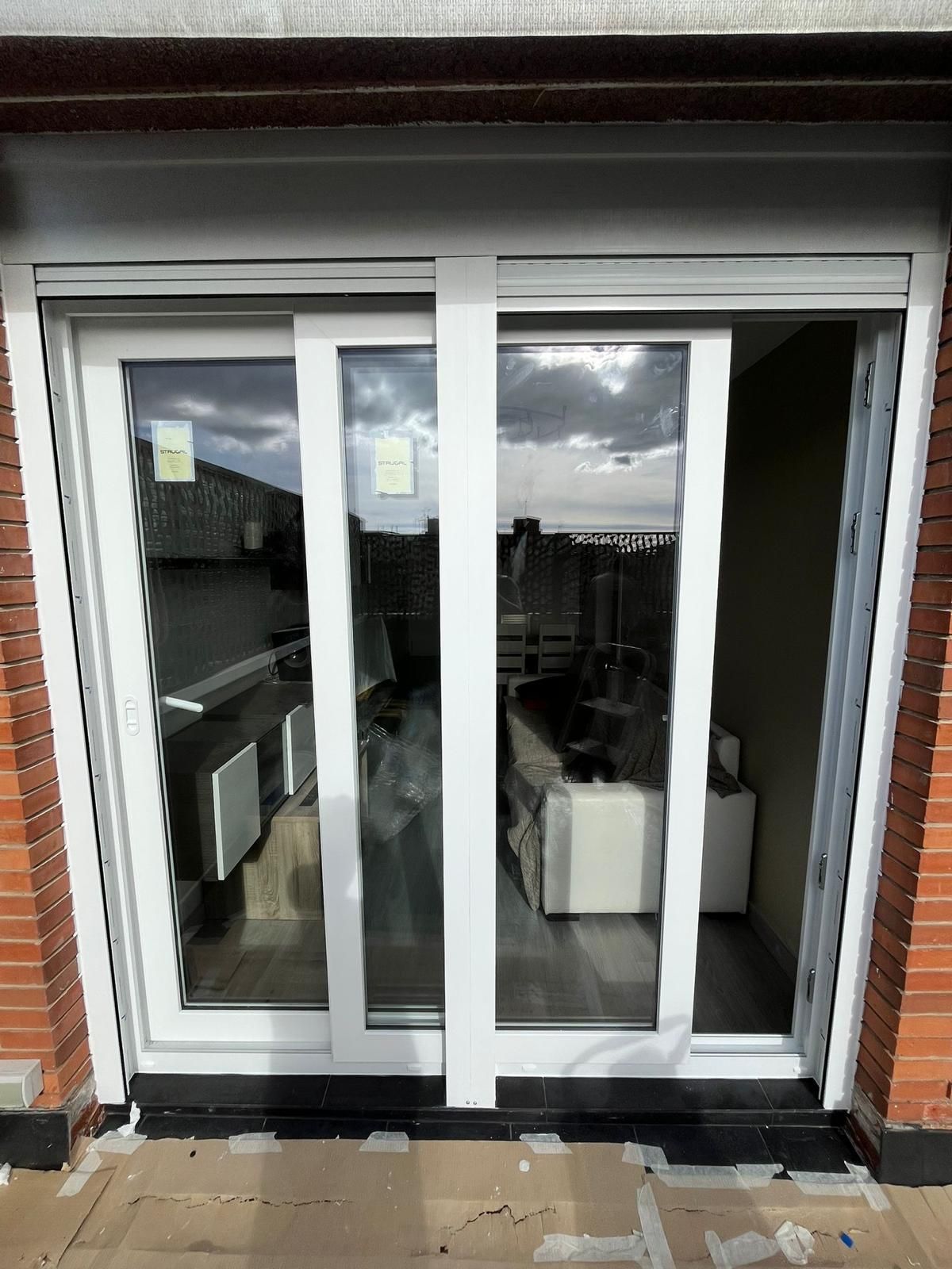 Ventana rota junto a una casa con una mosquitera doblada y plantas crecidas a lo largo de los cimientos.