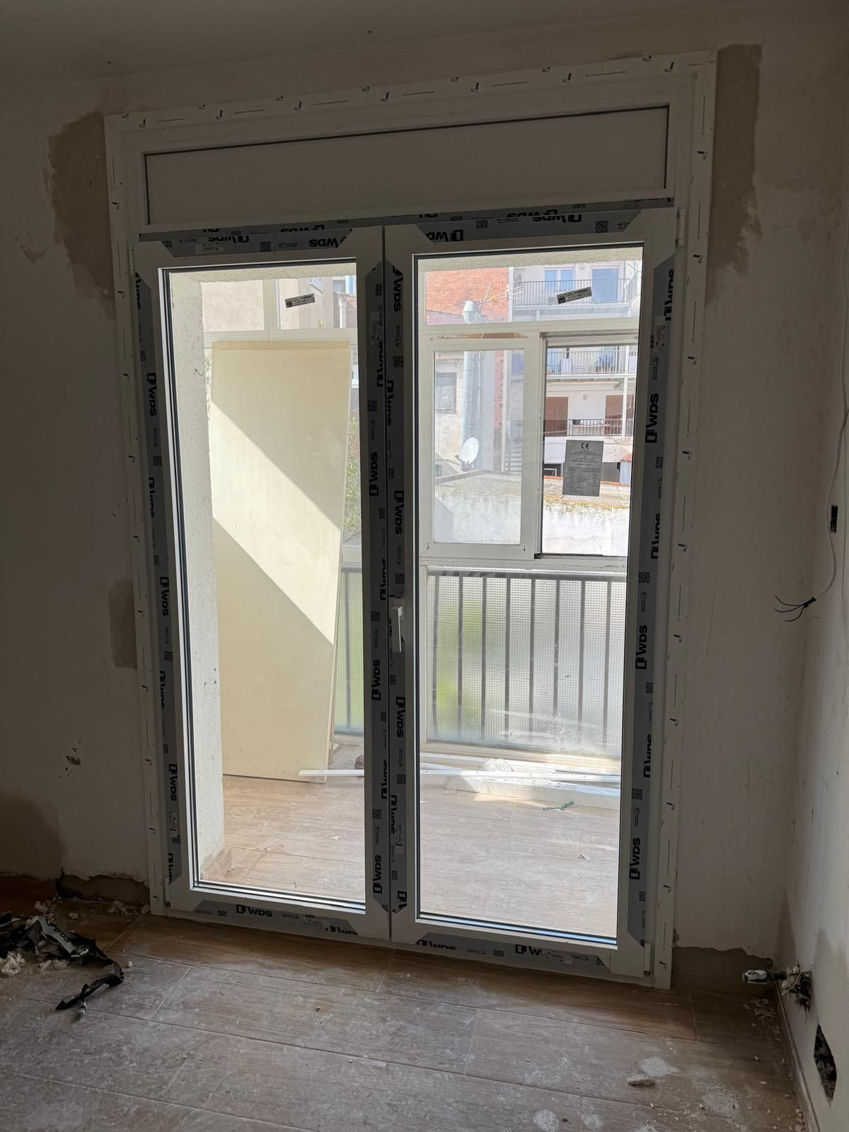 Habitación sin terminar con puertas de cristal con marcos blancos que dan a un balcón con vistas a la ciudad.