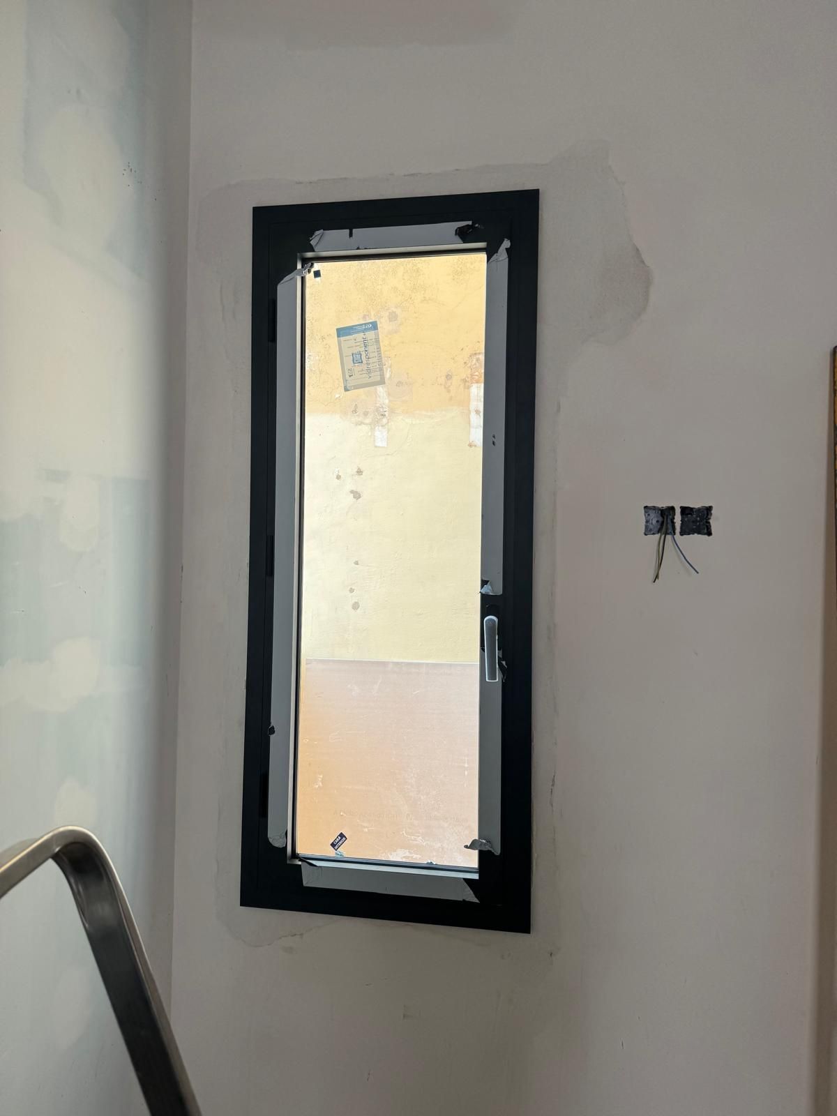 Una estrecha ventana con marco negro en una habitación blanca y desnuda, con cables eléctricos expuestos a su lado.