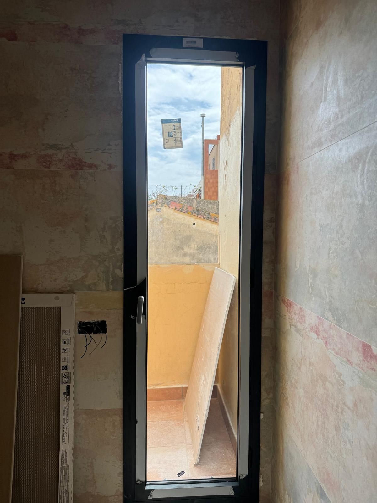 Entrada abierta a un balcón soleado, con una estrecha puerta de cristal y paredes interiores sin terminar.