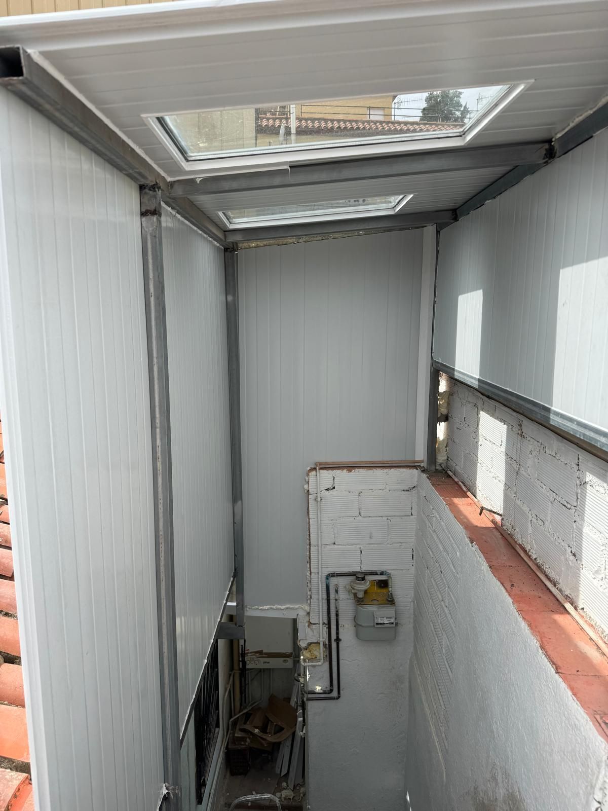 Estrecho pozo de acceso a la azotea con paredes metálicas, tragaluces en la parte superior y una escalera que desciende hacia abajo.