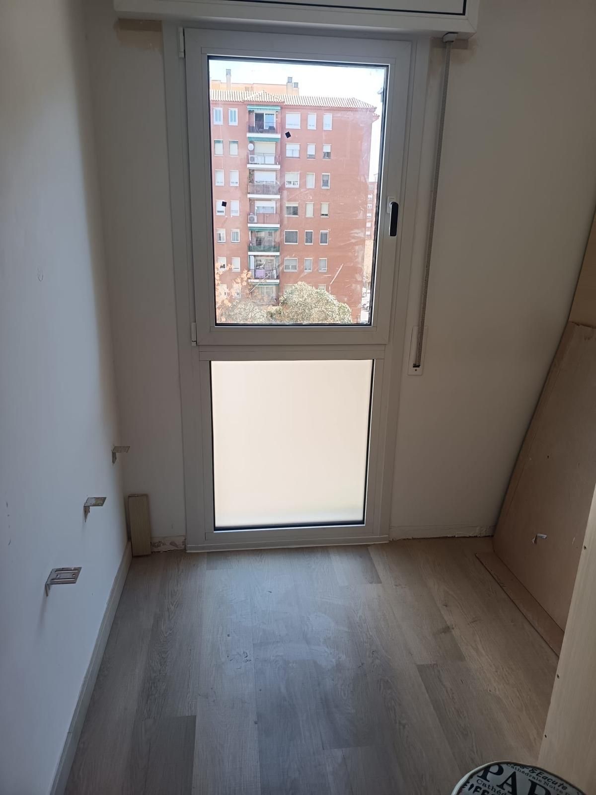Habitación estrecha y vacía con suelo de madera y una puerta con ventana que da a un edificio de apartamentos.
