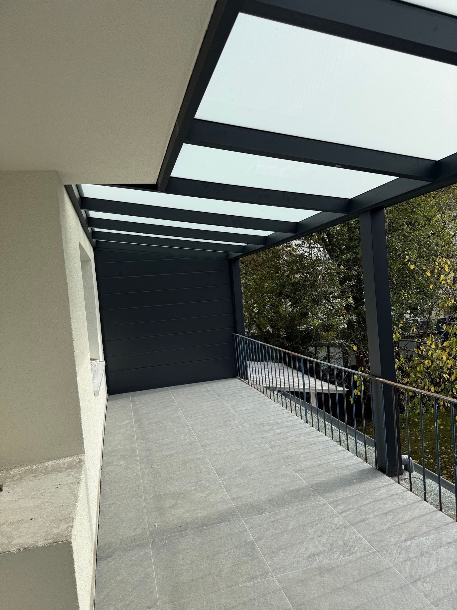 Ein moderner Balkon mit grau gefliestem Boden, schwarzem Metallrahmendach und Blick auf Bäume.