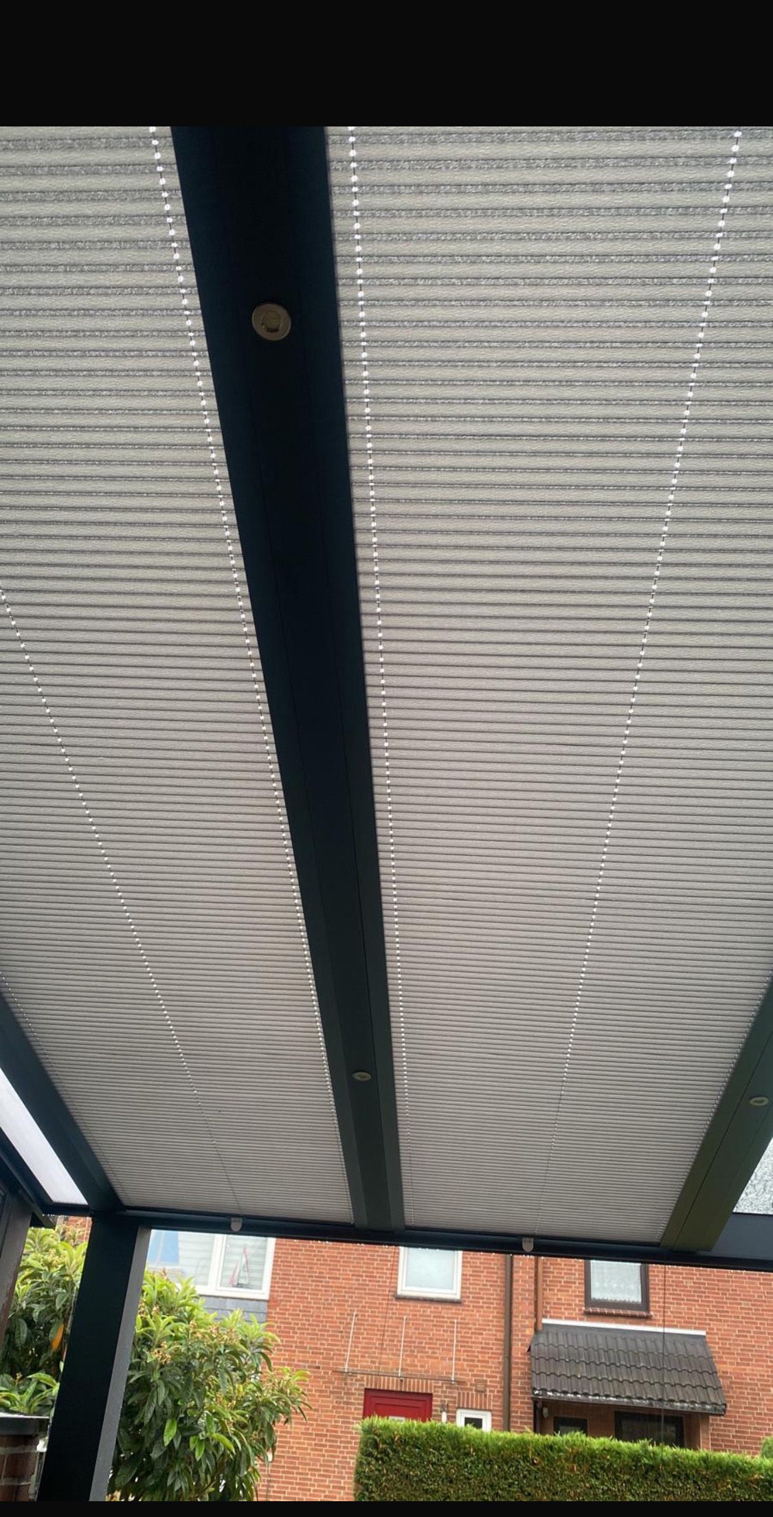 Blick auf ein Pergola-Dach mit verstellbaren grauen Lamellen über einem Backsteingebäude und grünen Bäumen.