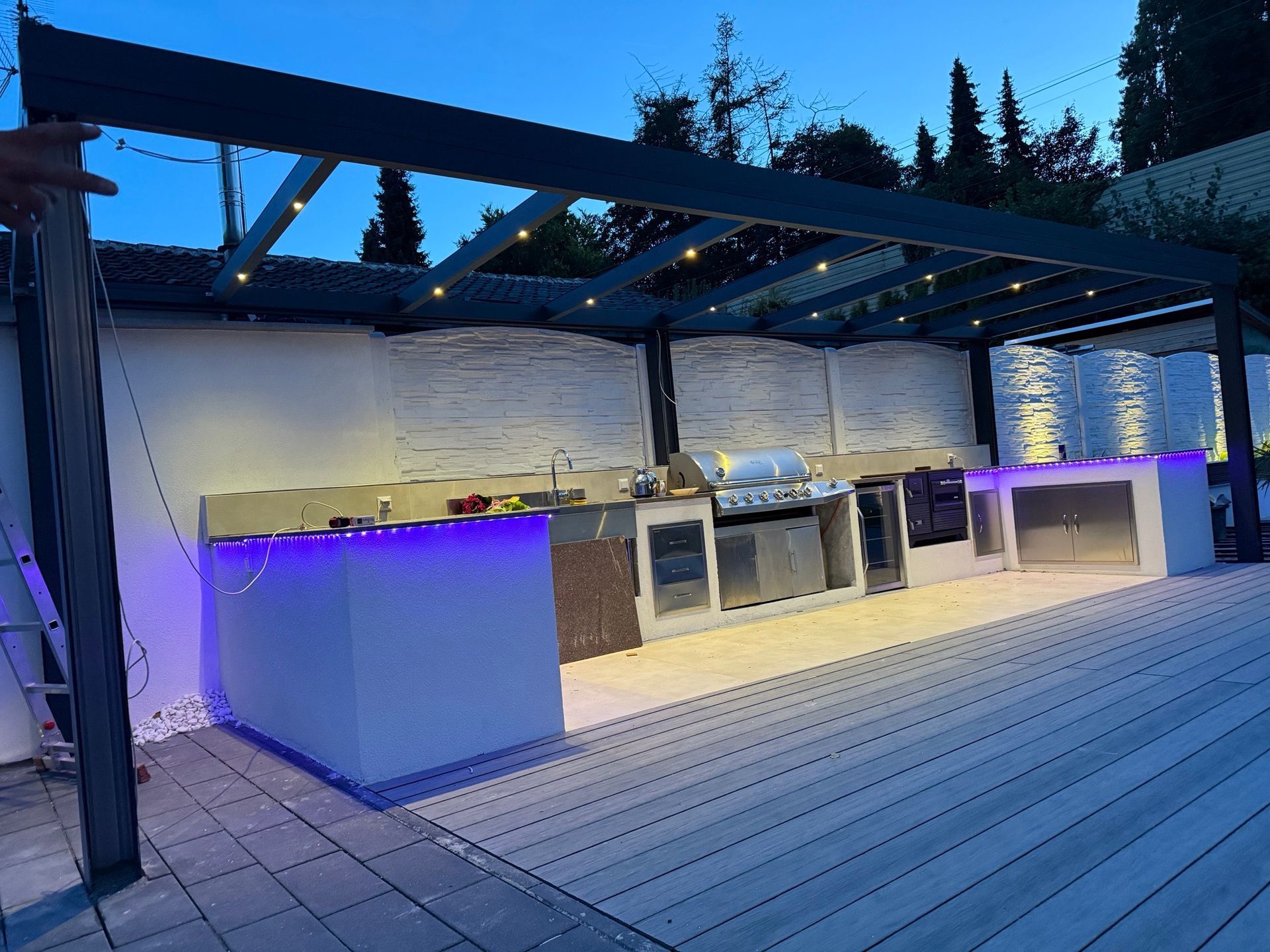 Außenküche mit beleuchteten Arbeitsflächen, Grill und überdachter Pergola in der Abenddämmerung.