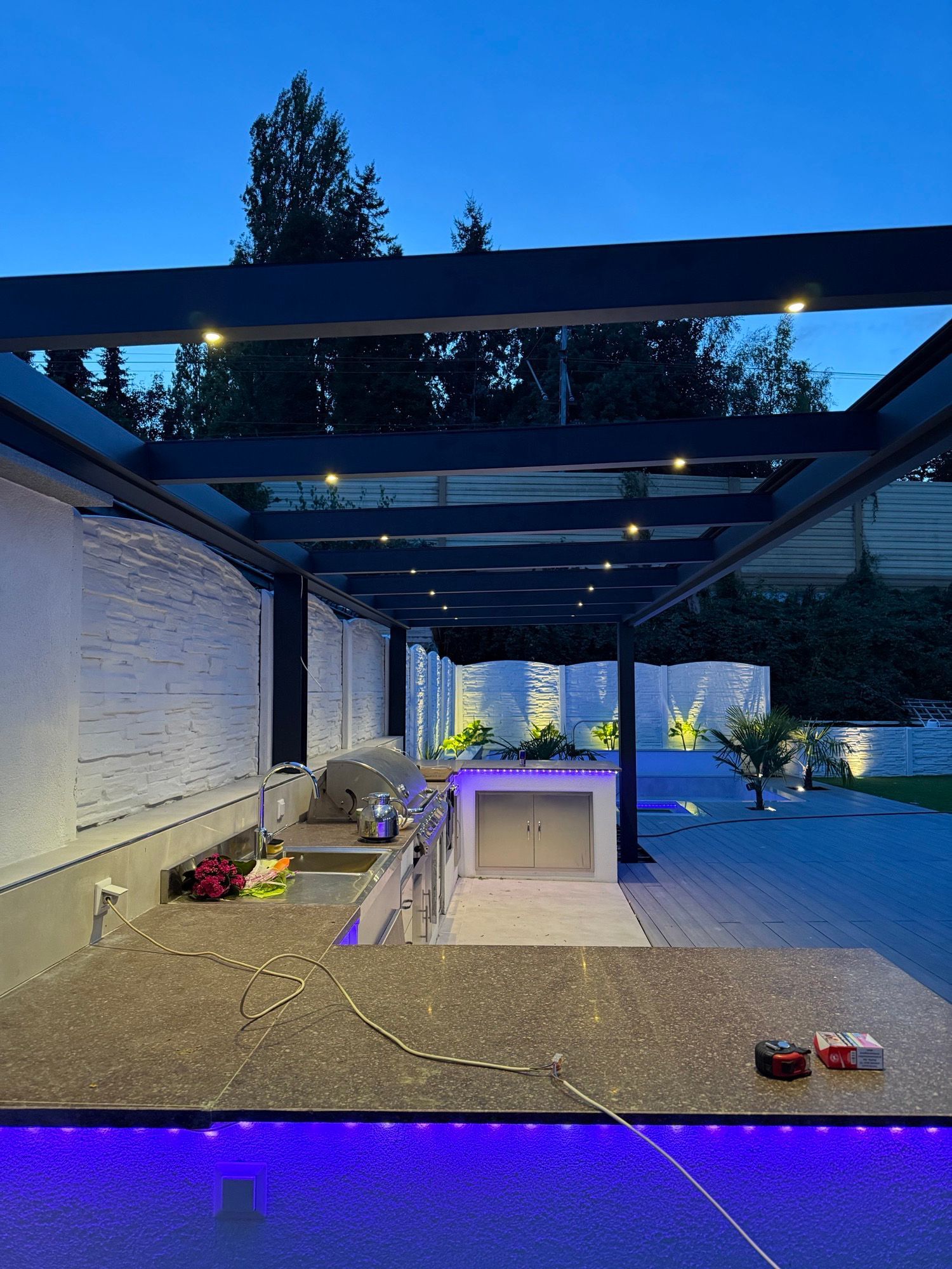 Außenküche mit dunkler Pergola, Arbeitsplatte mit blauen Lichtern und Abendhimmel.