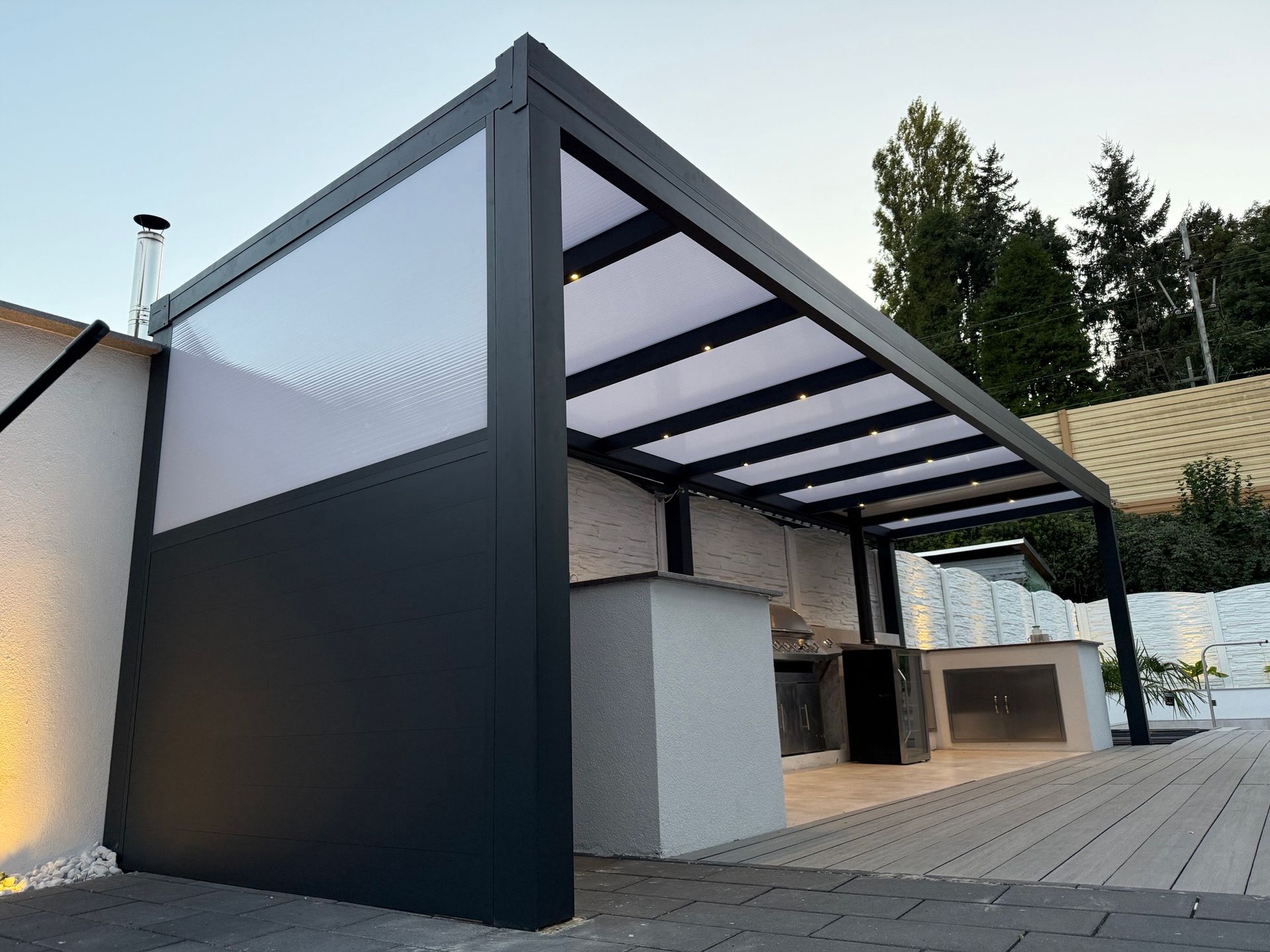 Moderne Außenküche mit dunkelgrauer Pergola, weißen Dachpaneelen und einer teilweise geschlossenen Seite.