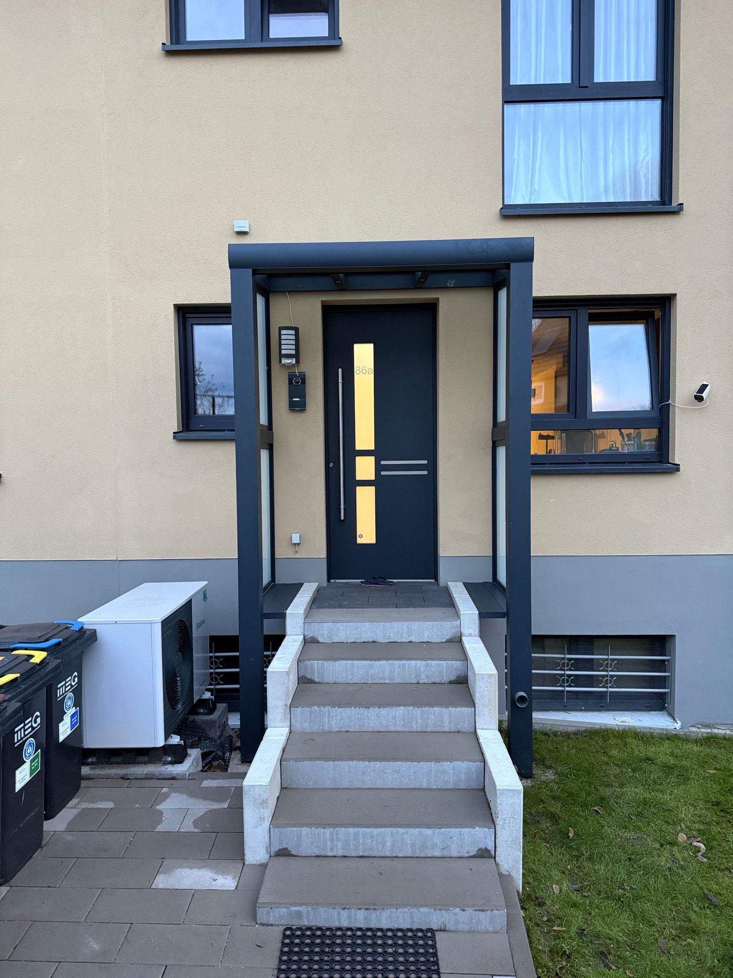 Grauer Eingang mit Stufen, die zu einer dunklen Tür unter einem Vordach führen. Fenster auf beiden Seiten.