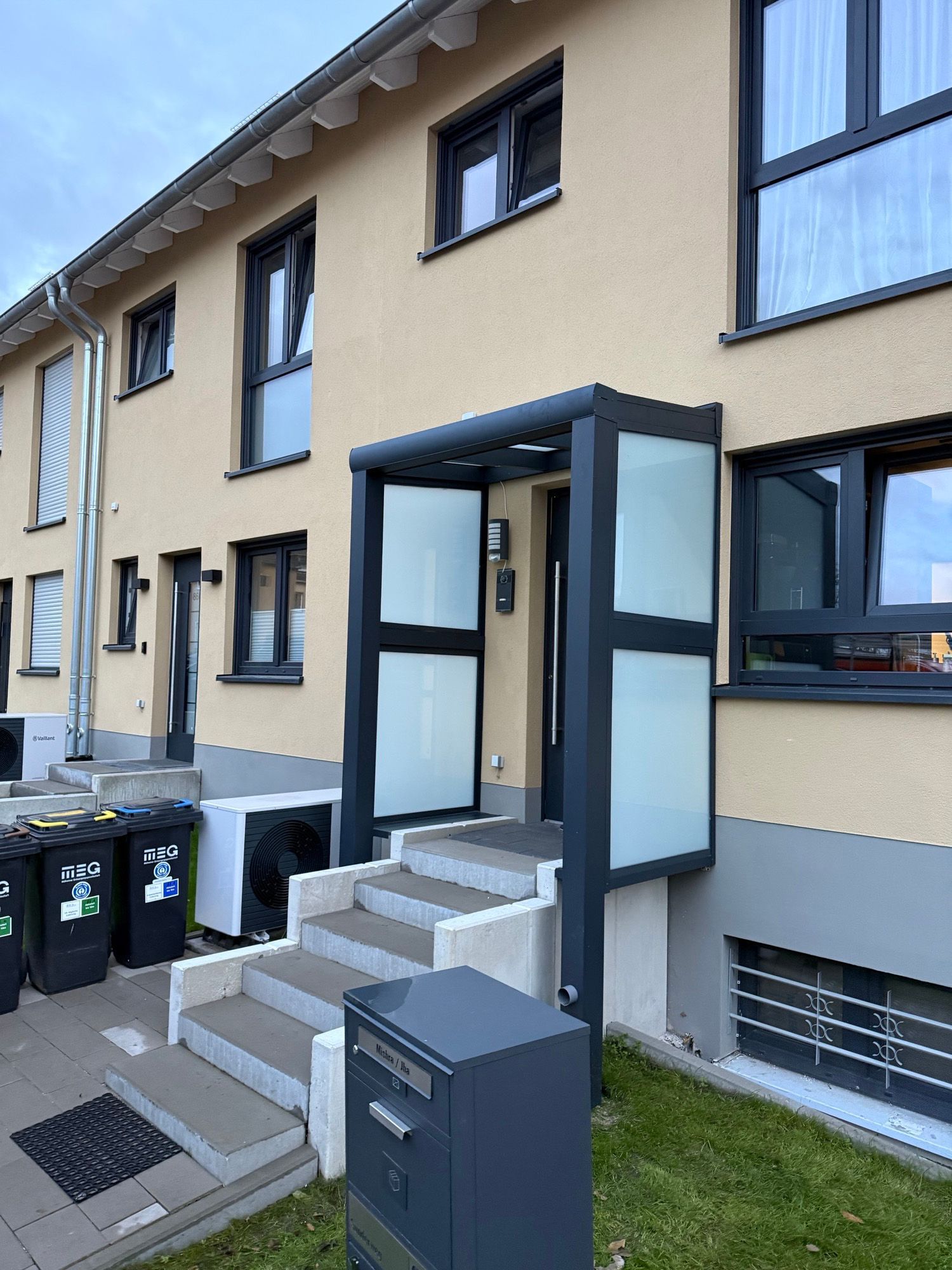 Fassade eines Reihenhauses mit grauem Eingangsvordach über den Stufen, die zur Haustür führen.