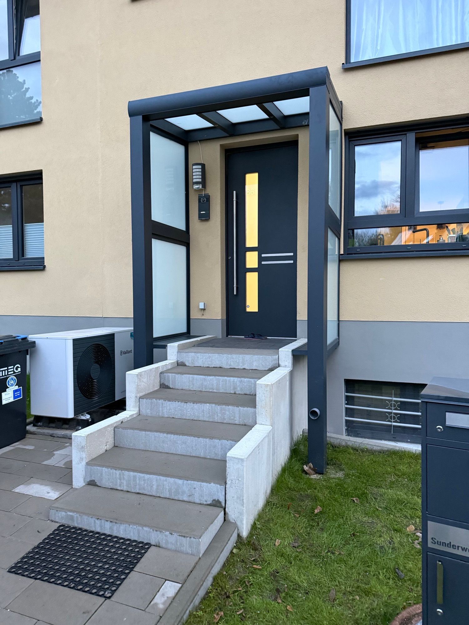 Graue Tür mit gelbem Streifen, überdachter Eingang und Treppe.