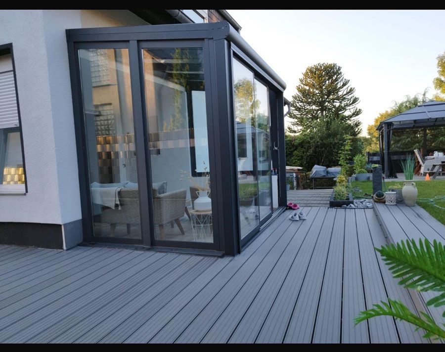 Ein moderner Wintergarten mit Glaswänden und Schiebetüren grenzt an eine graue Verbundholzterrasse mit Blick auf den Garten mit einem Pavillon.