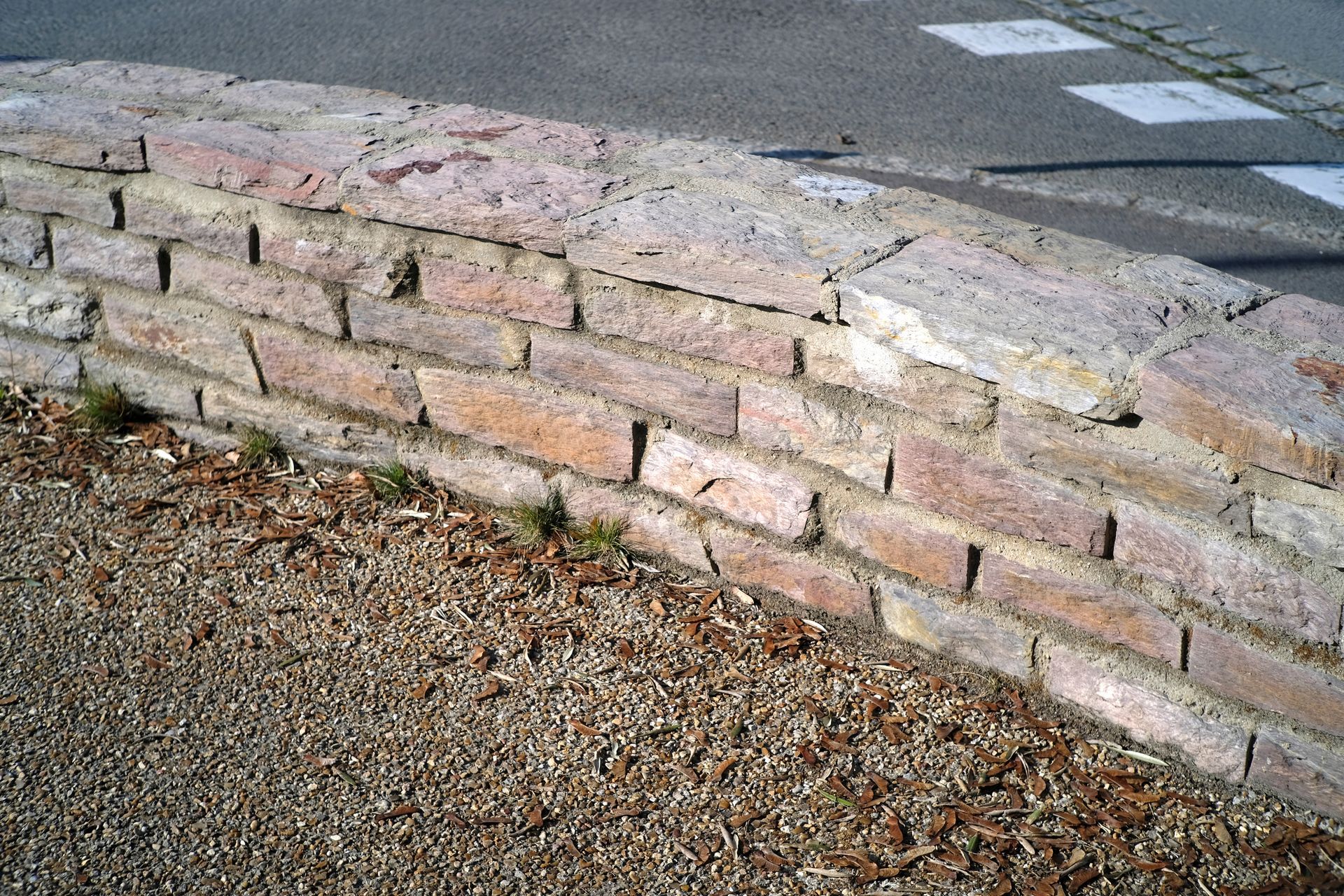 Mur de soutènement incurvé en pierre, bordant un chemin de gravier et une route pavée.