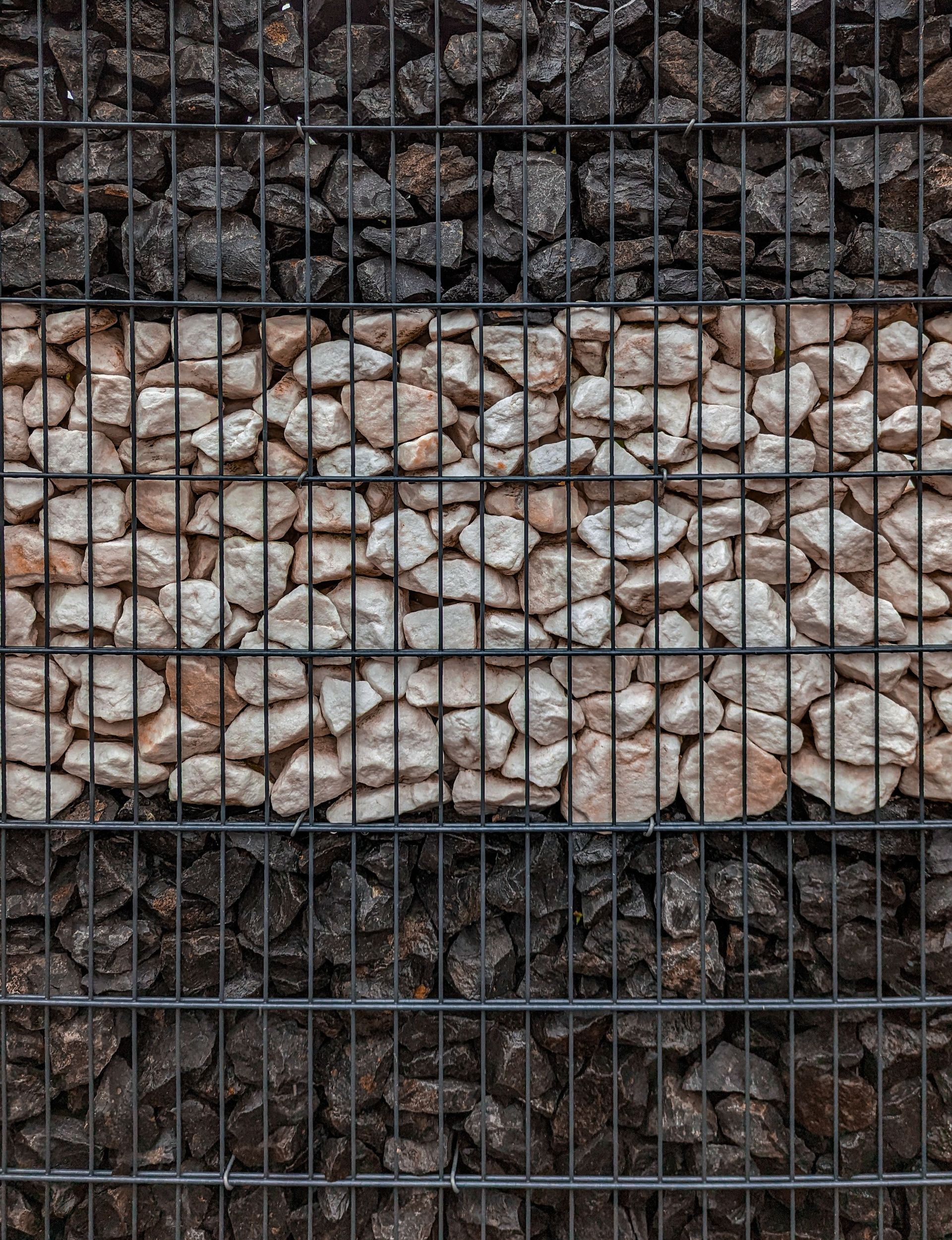 Pierres noires et beiges empilées, maintenues dans un mur de gabions en treillis métallique.