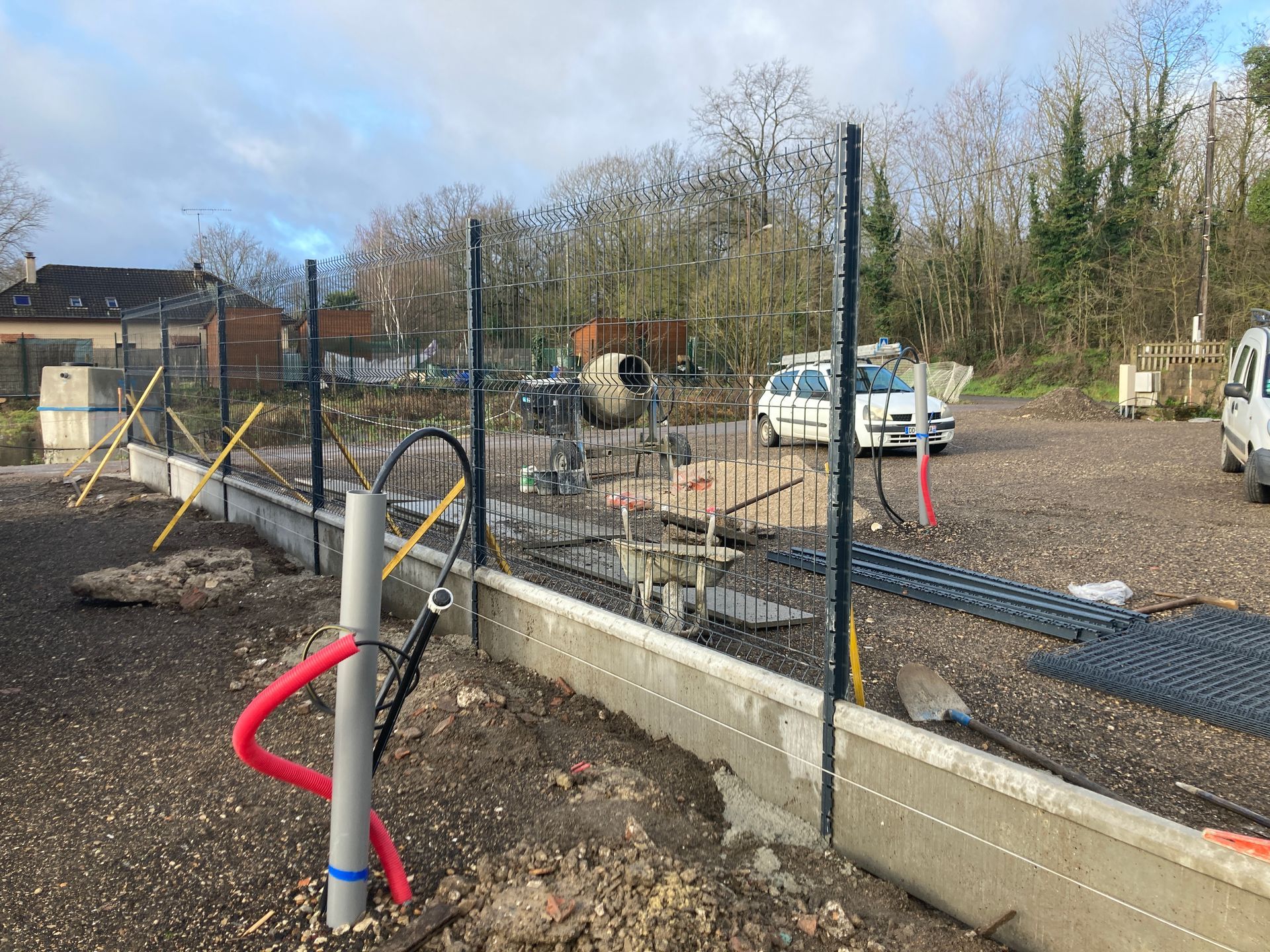 Chantier avec dalle de béton, poteaux de clôture métalliques et conduits électriques.
