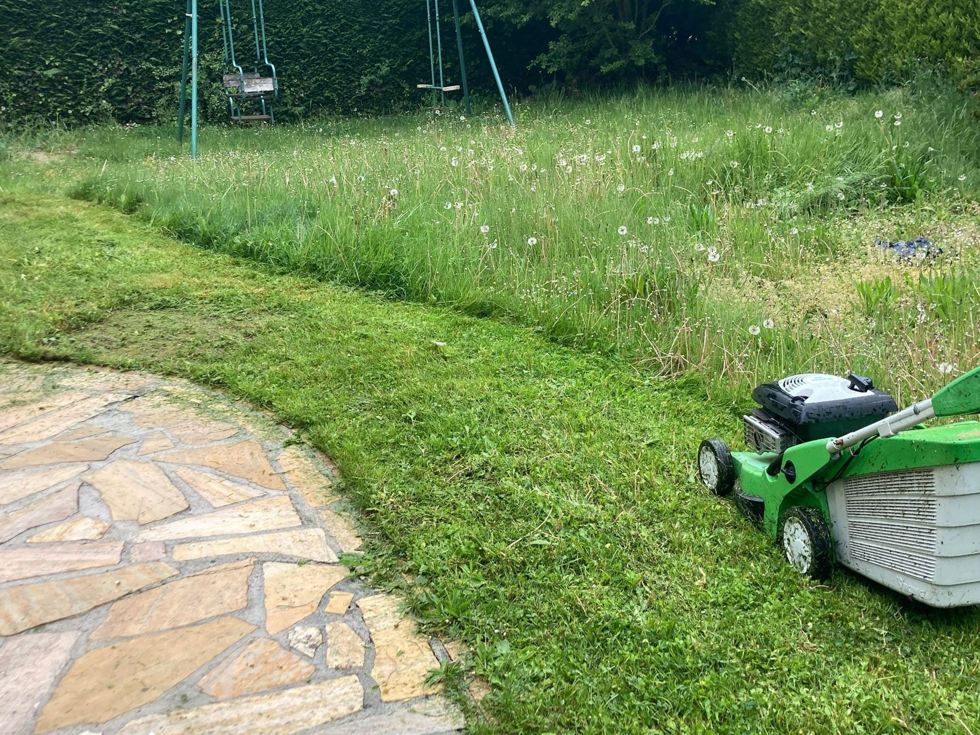 Une tondeuse à gazon verte coupe l'herbe dans un jardin avec une balançoire.