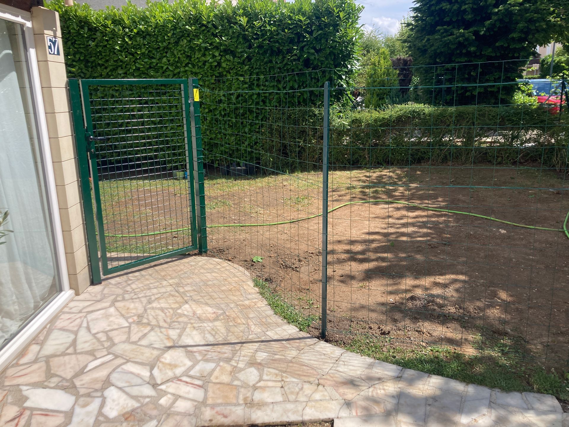 Portail et clôture verts donnant sur une cour arrière gazonnée avec terrasse en pierre, entourée de haies.