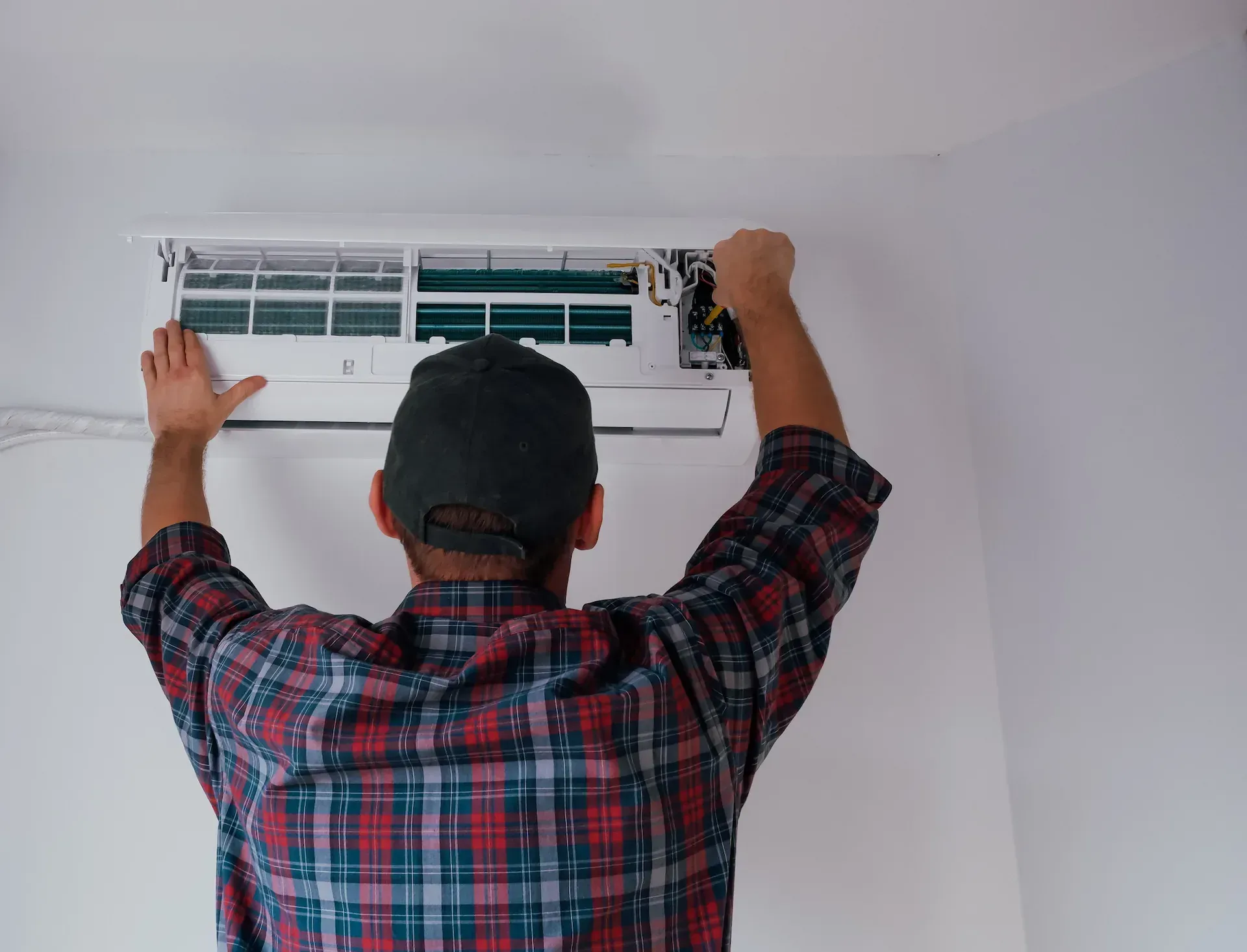 Una persona con una camisa a cuadros está instalando una unidad de aire acondicionado en una pared blanca.