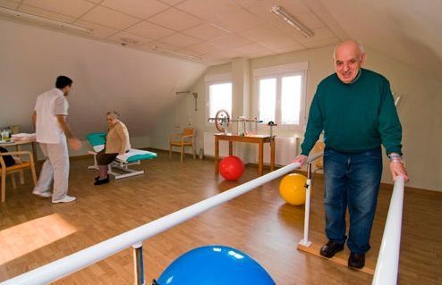 Un hombre mayor camina sobre barras paralelas en un gimnasio.