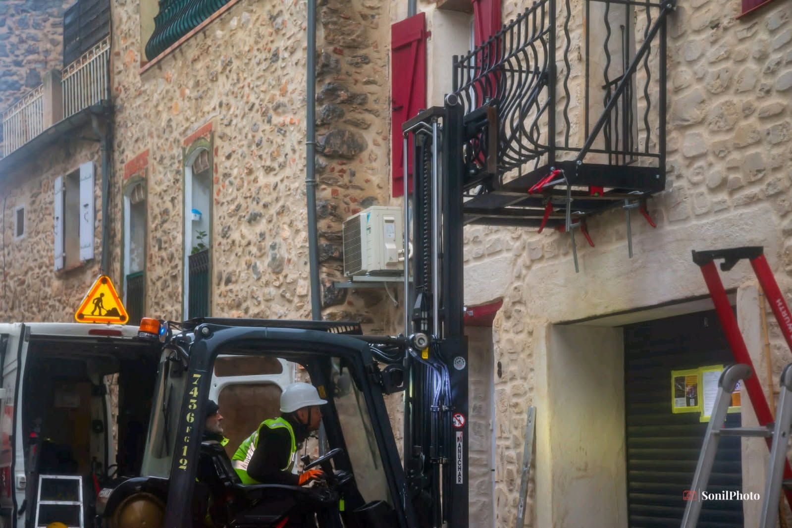 Un chariot élévateur soulève un balcon noir pour le mettre en place sur un bâtiment en pierre.