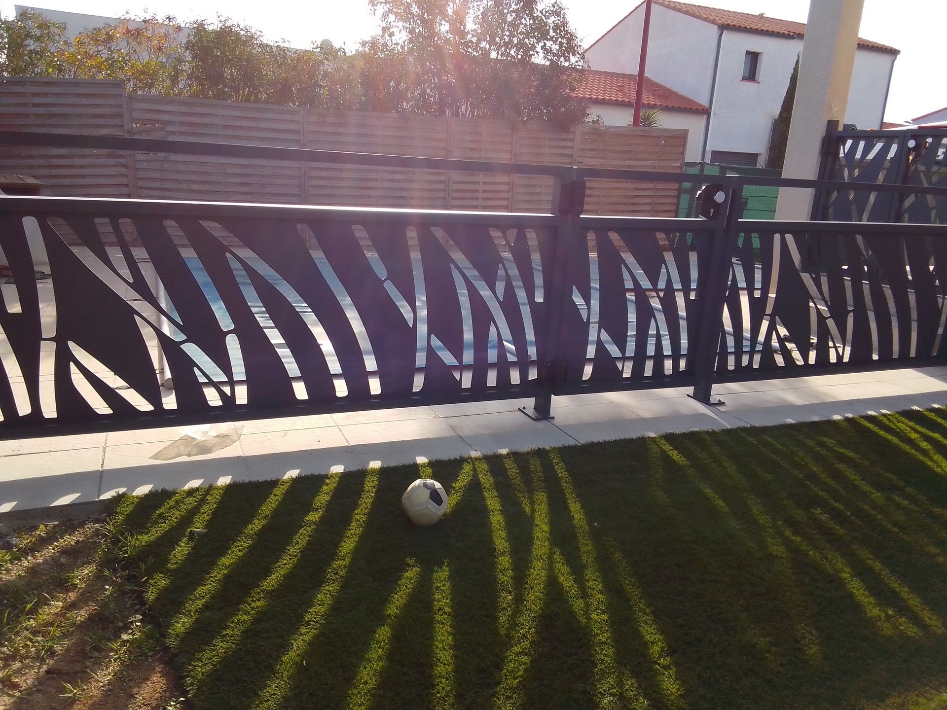 Clôture en métal noir avec un motif de feuille, projetant des ombres sur une pelouse verte.