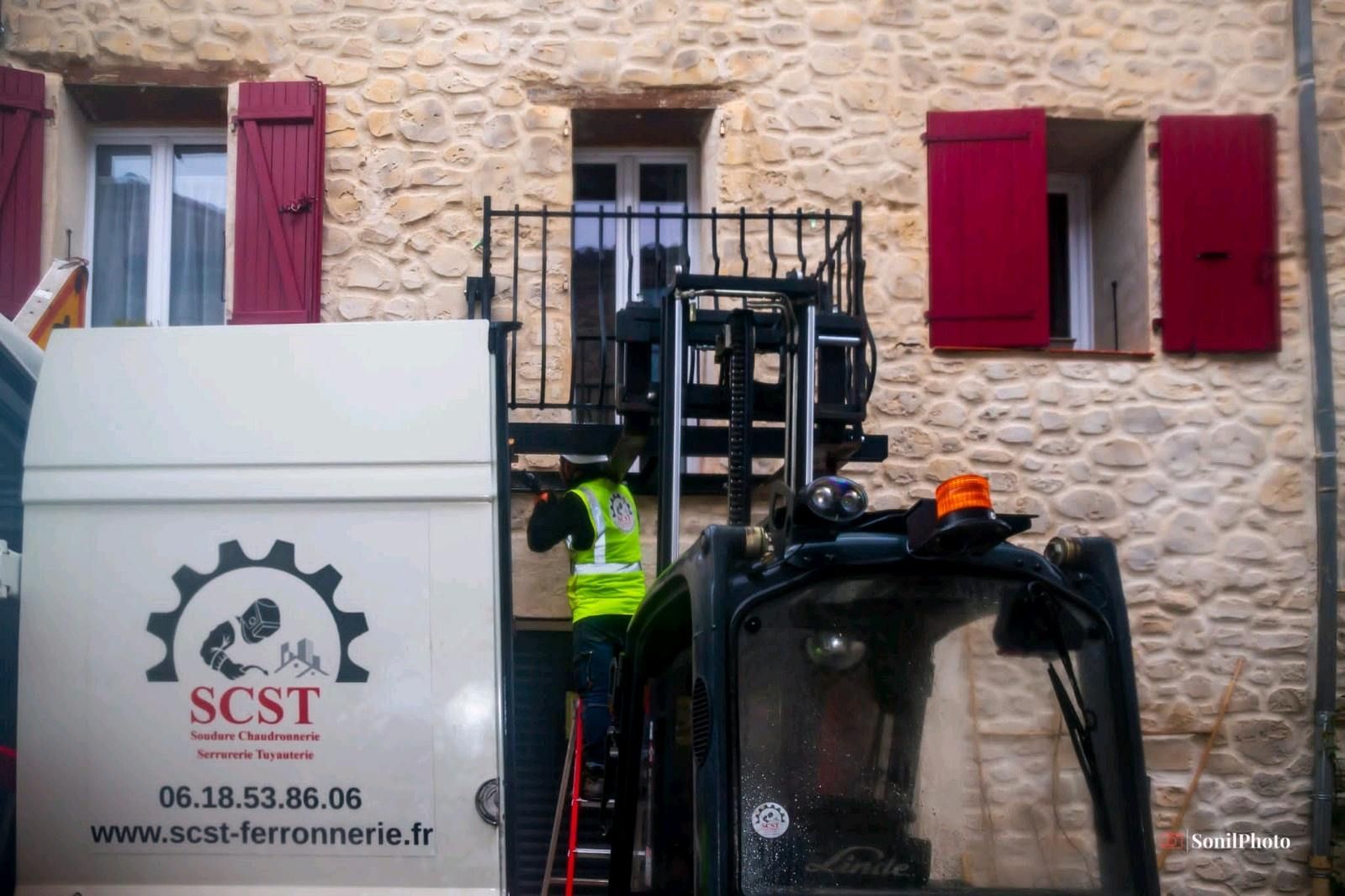 Un chariot élévateur avec un ouvrier près d'un bâtiment en pierre, déchargeant un camion portant le logo SCST.