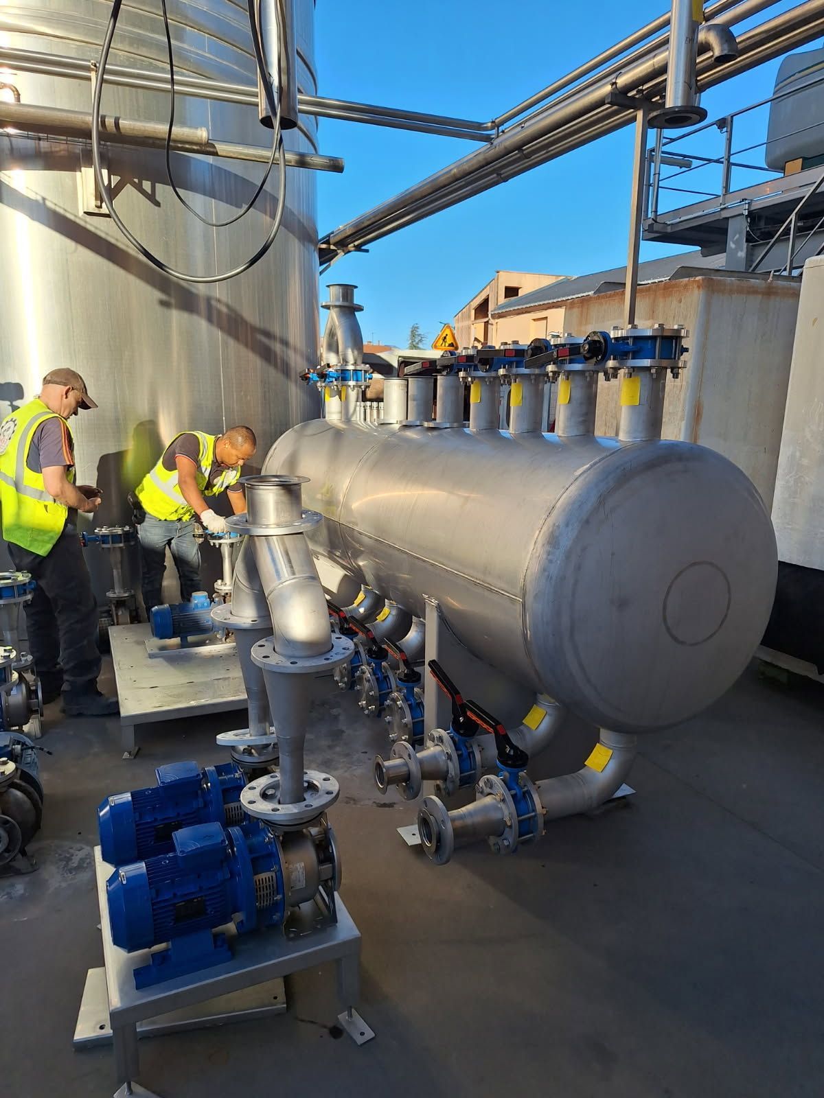 Deux ouvriers installent des équipements industriels : réservoirs, tuyaux et pompes à l'extérieur.