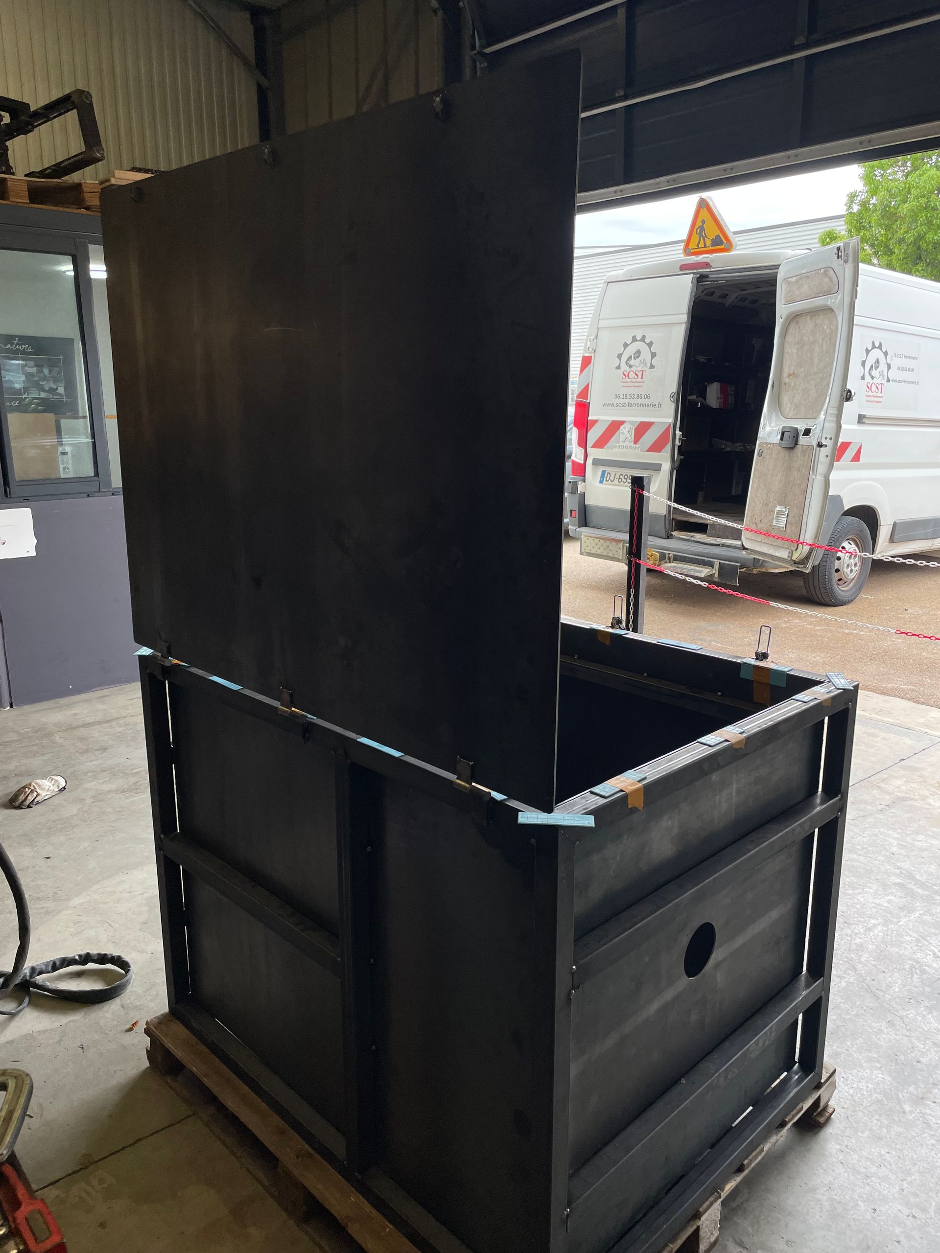 Boîte en métal noir avec couvercle ouvert, possiblement industrielle, à l'intérieur d'un atelier.